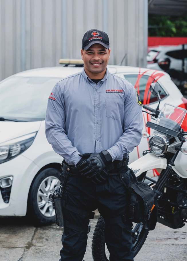 Security guard in uniform with cap and gloves standing in front of a white car and a motorcycle.