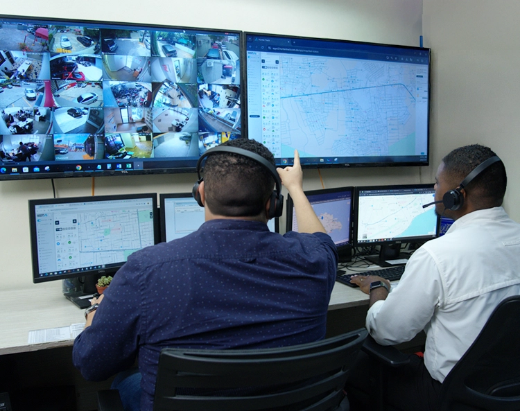 Two security operators wearing headsets monitoring multiple surveillance camera feeds and maps on large screens in a control room.