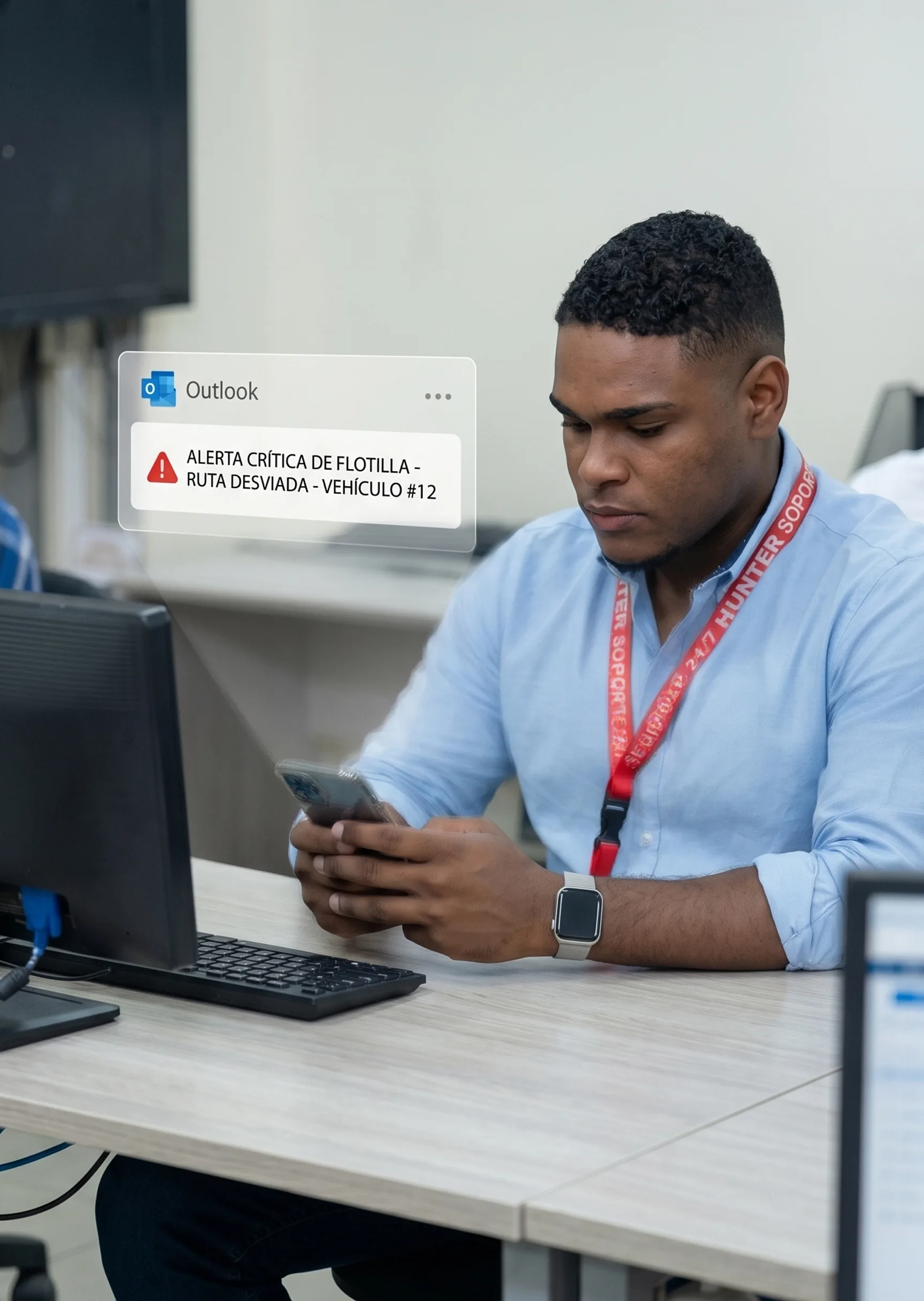 Man in blue shirt sitting at a desk, holding a smartphone, with a digital fleet alert message displayed near him.