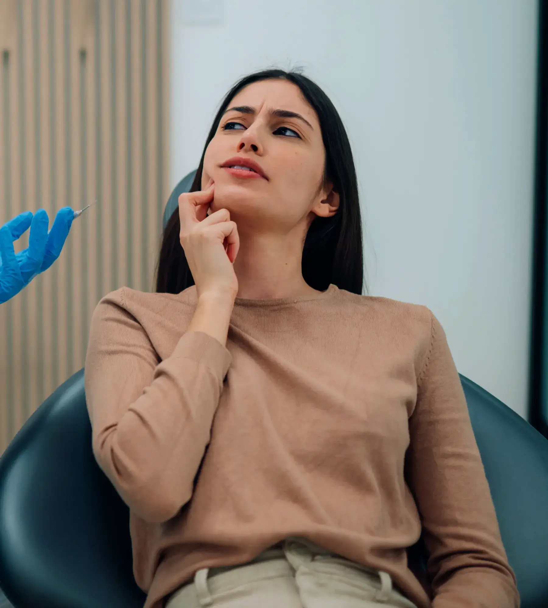 A woman sits in a chair, looking uncertain, while a gloved hand holds a needle nearby.