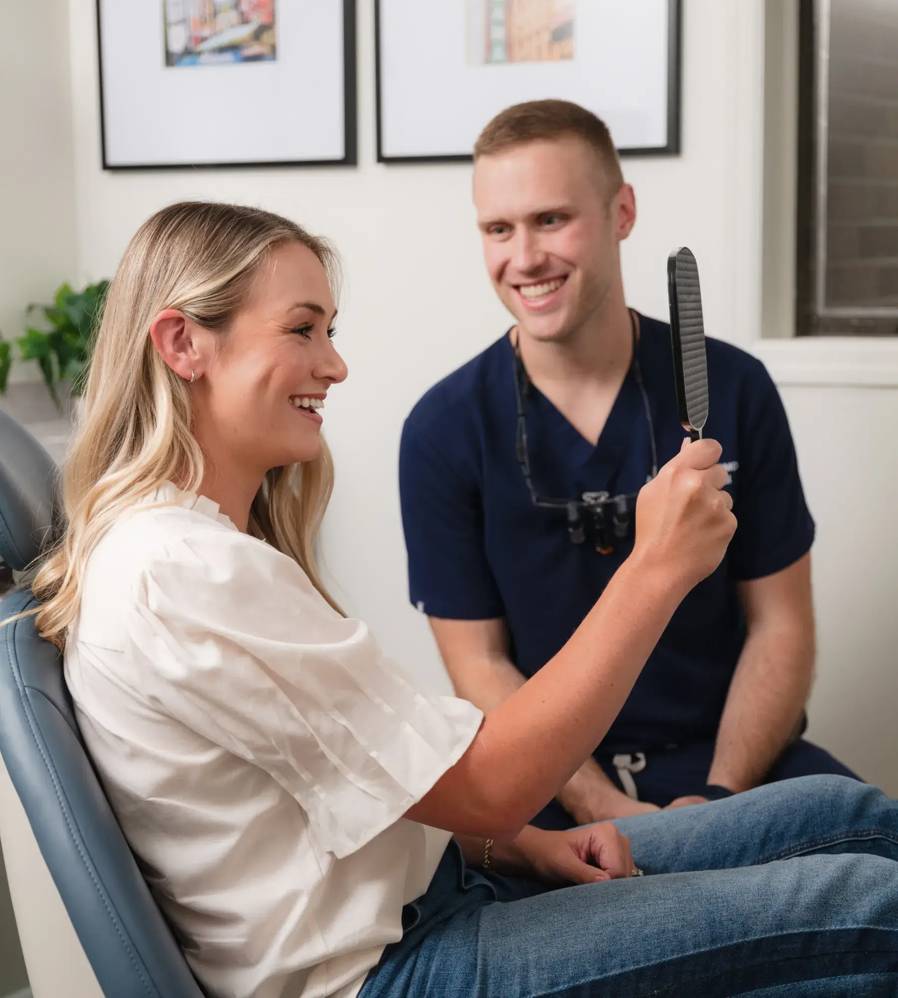 A woman sits in a dental chair, smiling while holding a hand mirror, with a dentist nearby.