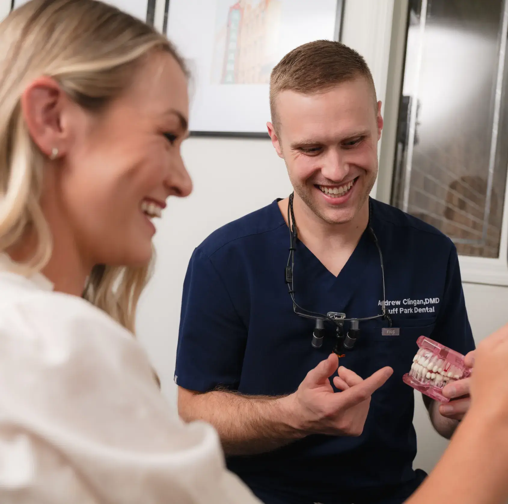 A woman sits in a dental chair smiling into a handheld mirror, with a dentist nearby.