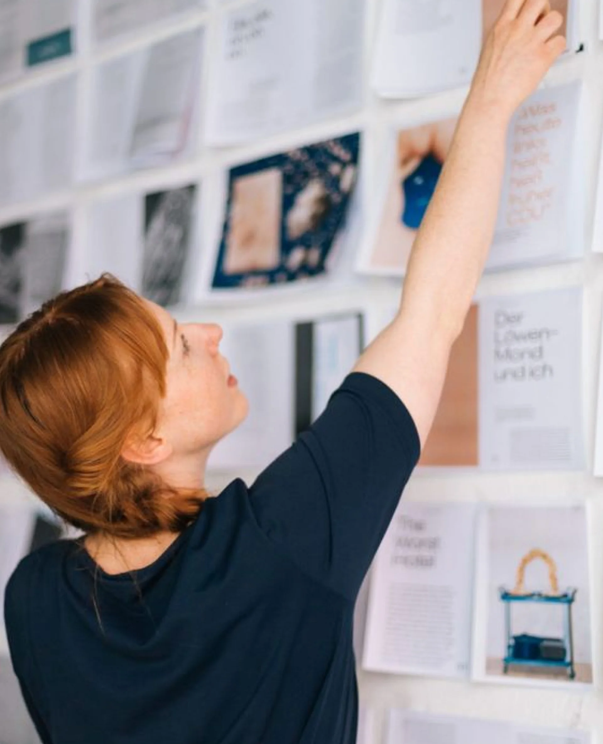 Woman with red hair in a braid reaching up to adjust papers on a wall covered with various documents and images.