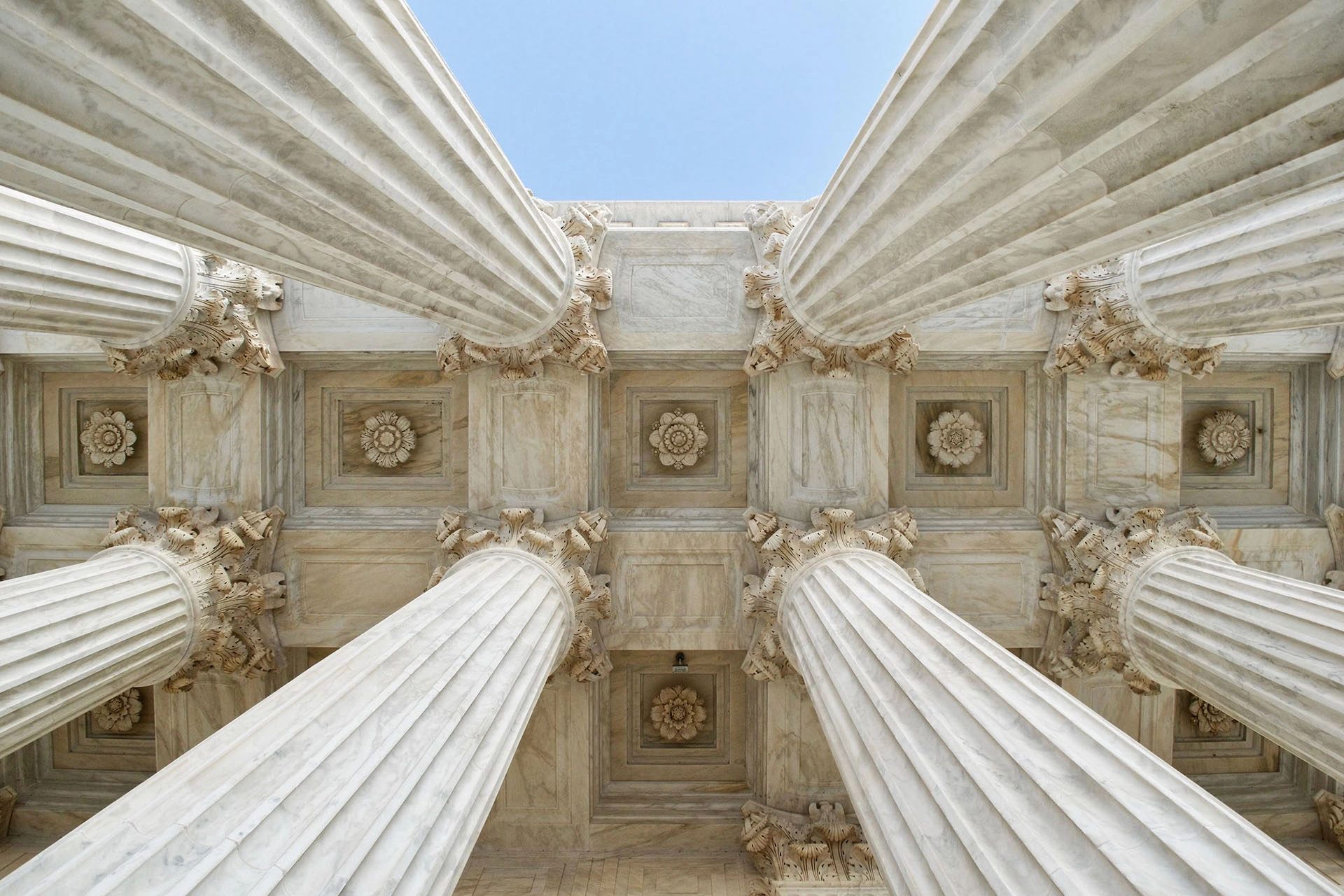 Upward view of classical marble columns with ornate capitals and decorated ceiling panels under a clear sky.
