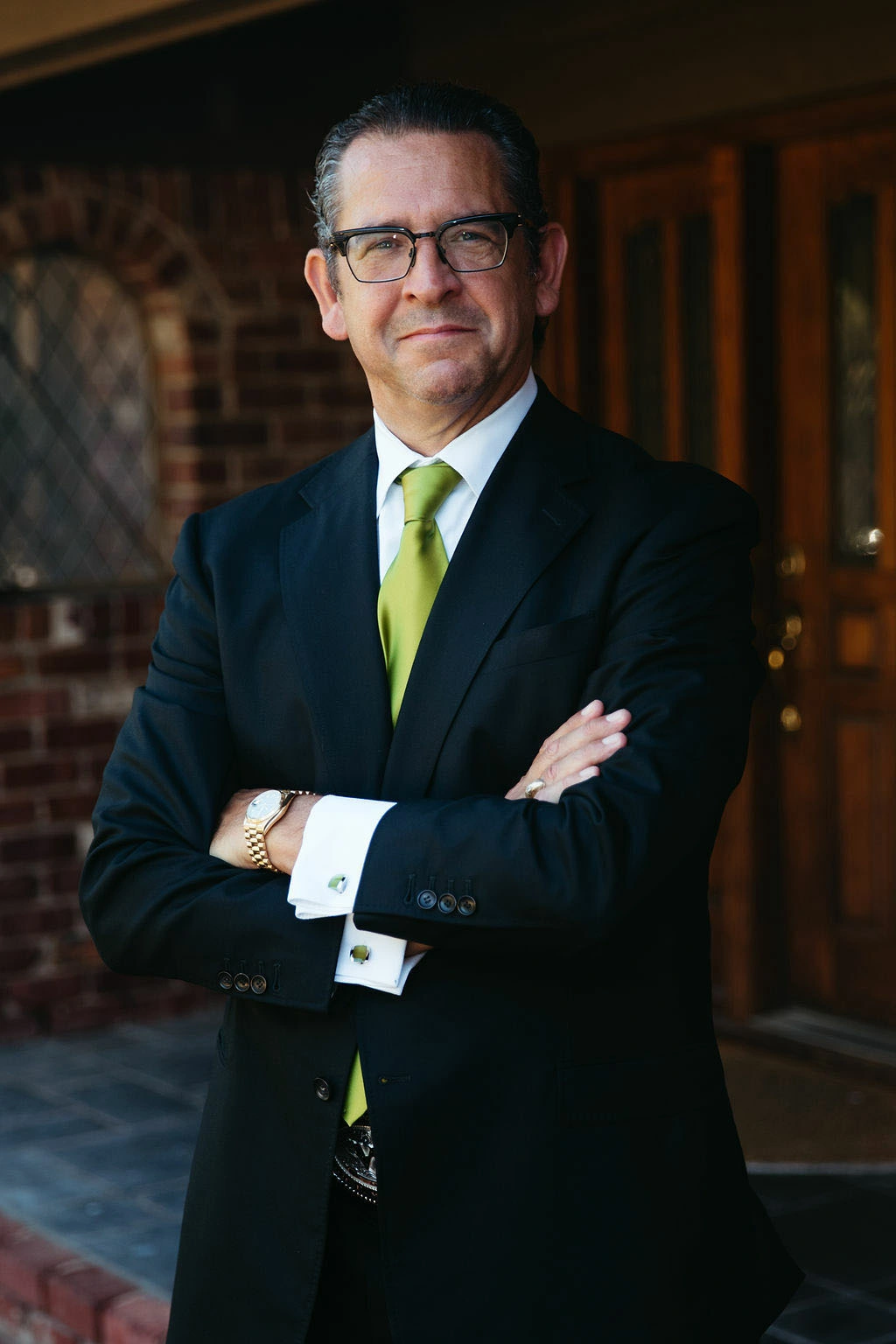 Man in glasses wearing a black suit with a white shirt and lime green tie, standing with arms crossed in front of a brick wall and wooden door.