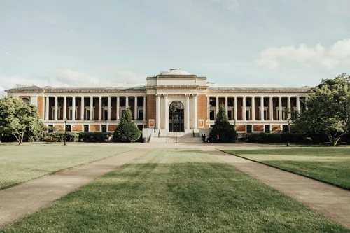 Wide view of a historic academic building with columns and a central dome, surrounded by green lawns and trees.