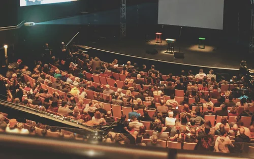 Audience seated in a theater facing a stage with two stools and a large screen.