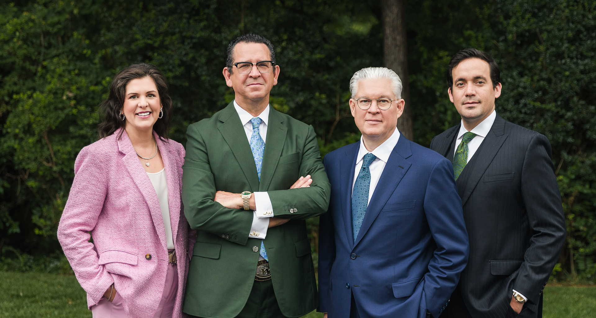 Four professionally dressed individuals standing outdoors in front of green foliage, with two men wearing suits and ties, one man in a green jacket, and one woman in a pink blazer.