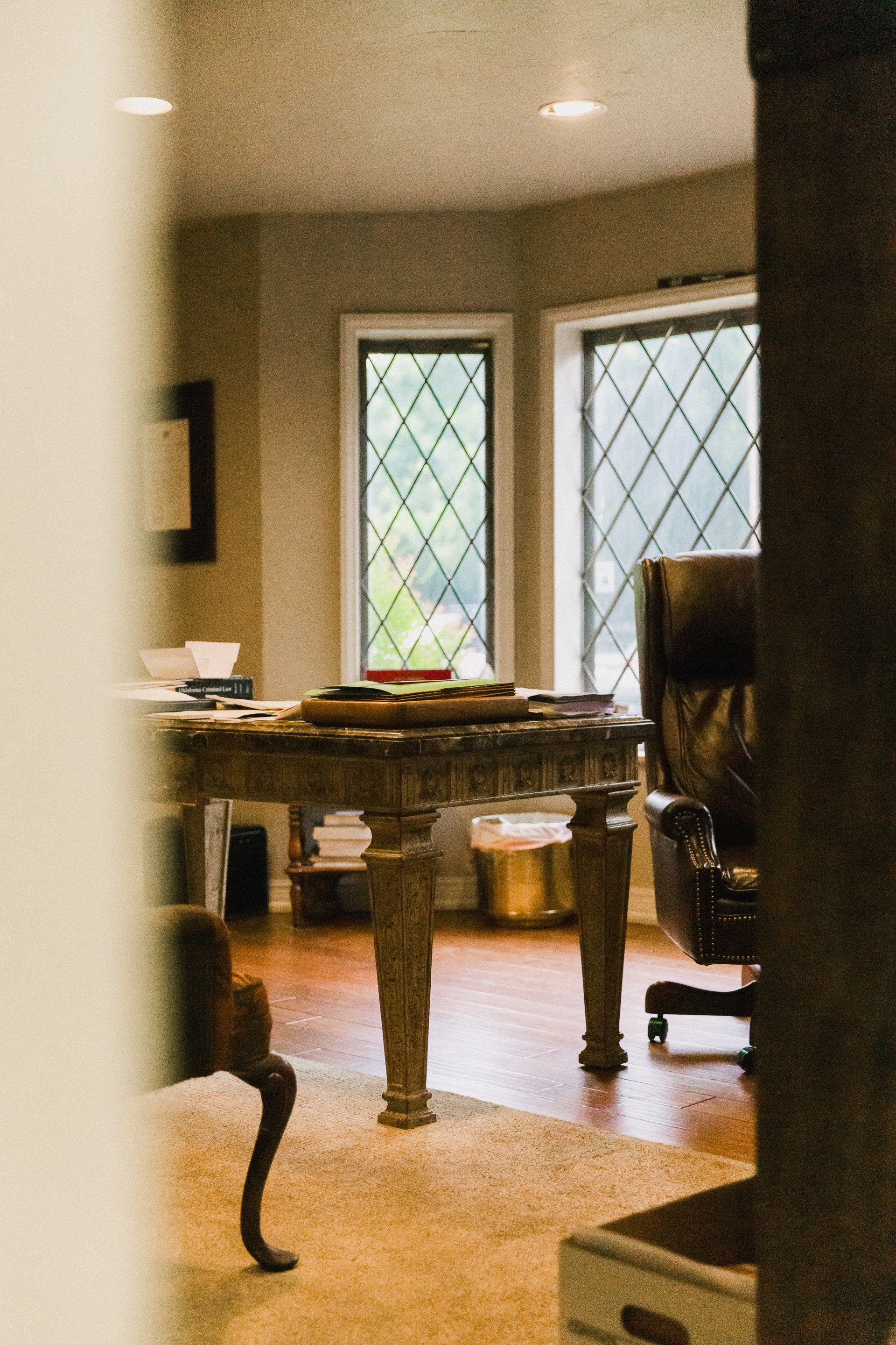 Wooden office desk with papers and books in front of diamond-patterned windows, alongside a leather office chair.