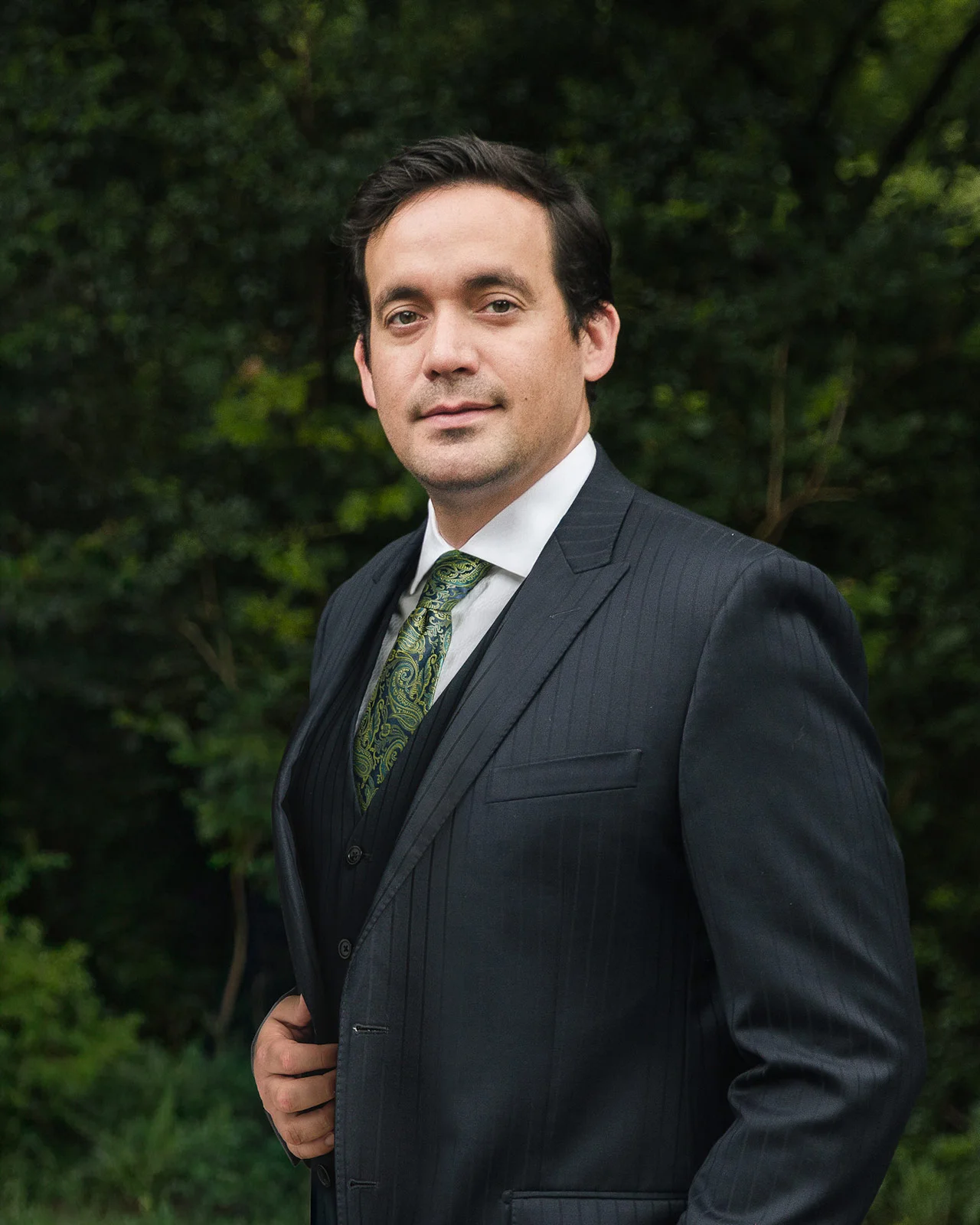 Man in a dark pinstripe suit with a green paisley tie standing outdoors with greenery in the background.