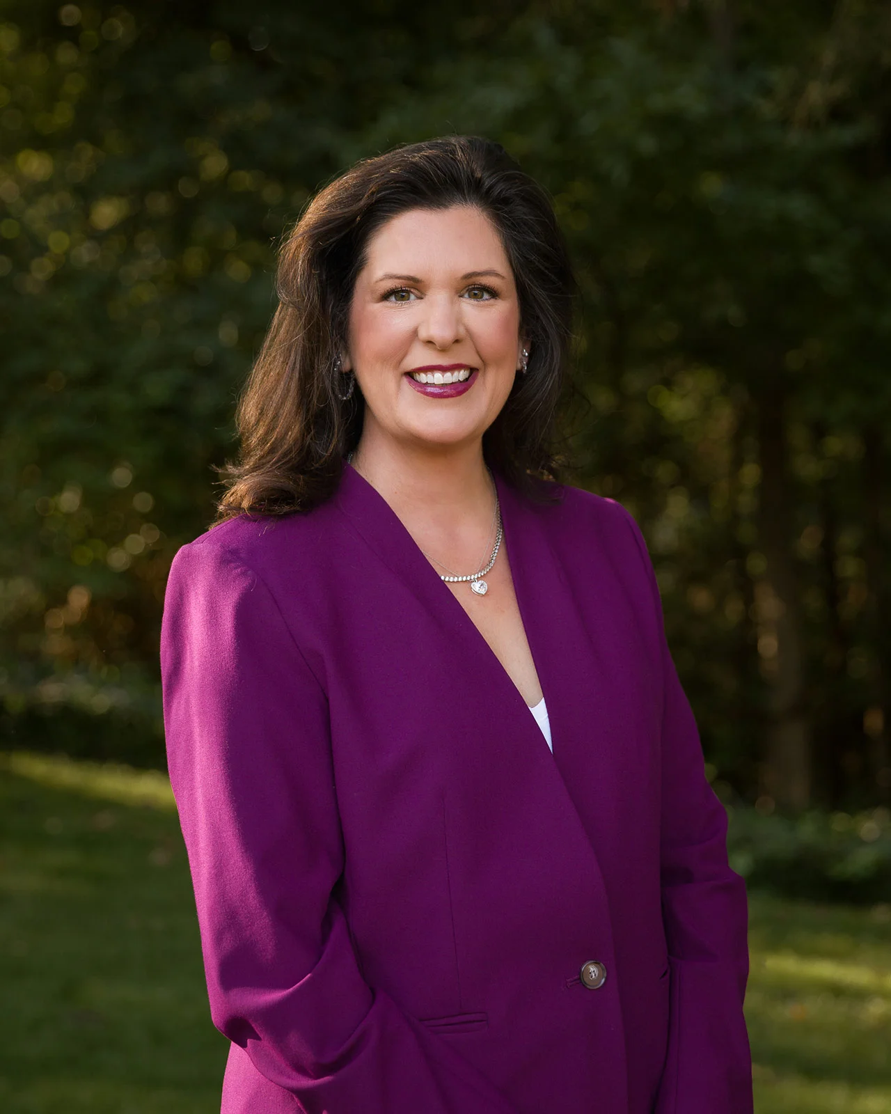 Smiling woman with dark hair wearing a purple blazer and silver necklace standing outdoors with green foliage background.