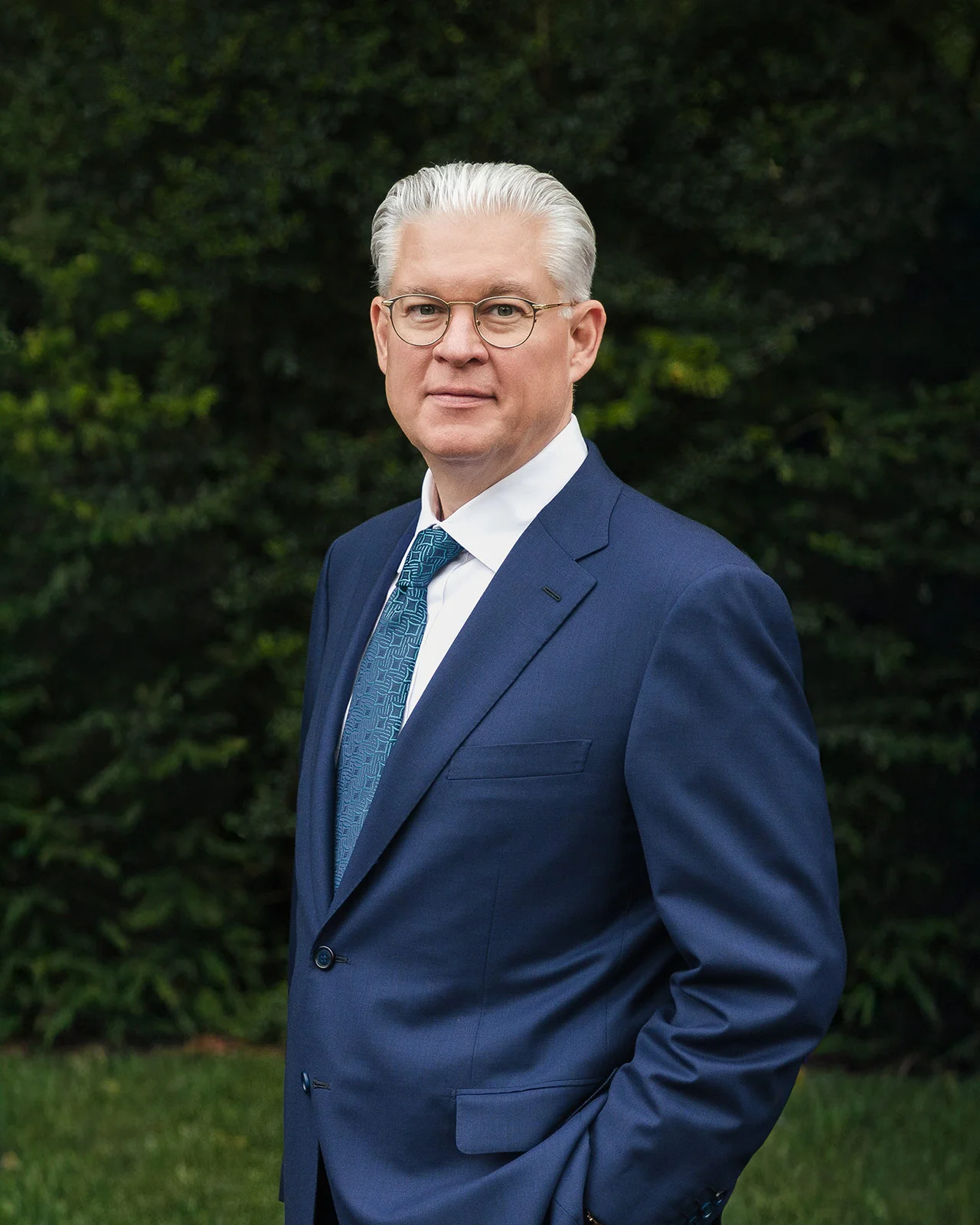 Mature man with white hair and glasses wearing a blue suit and patterned tie standing outdoors with greenery in the background.