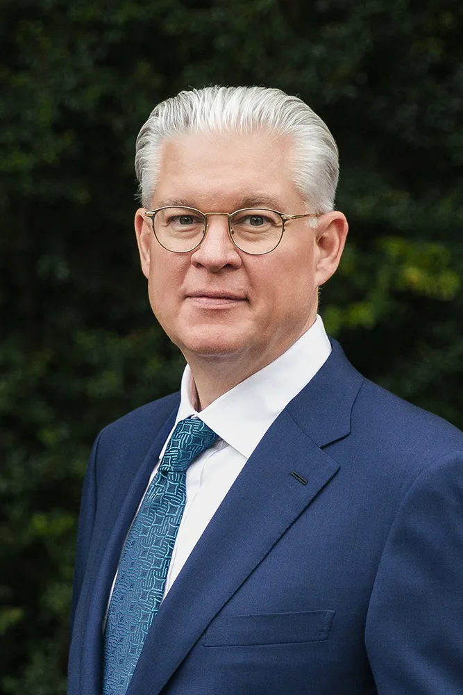Man with white hair and glasses wearing a blue suit and patterned blue tie, standing outdoors with green foliage background.