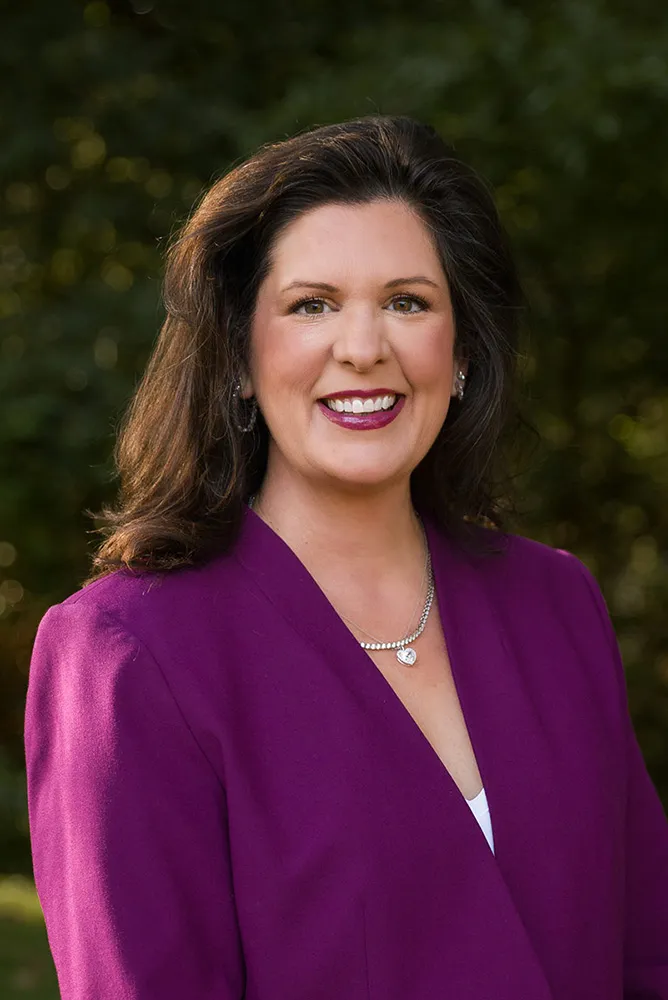 Smiling woman with shoulder-length brown hair wearing a purple blazer and a silver necklace, standing outdoors with blurred greenery background.