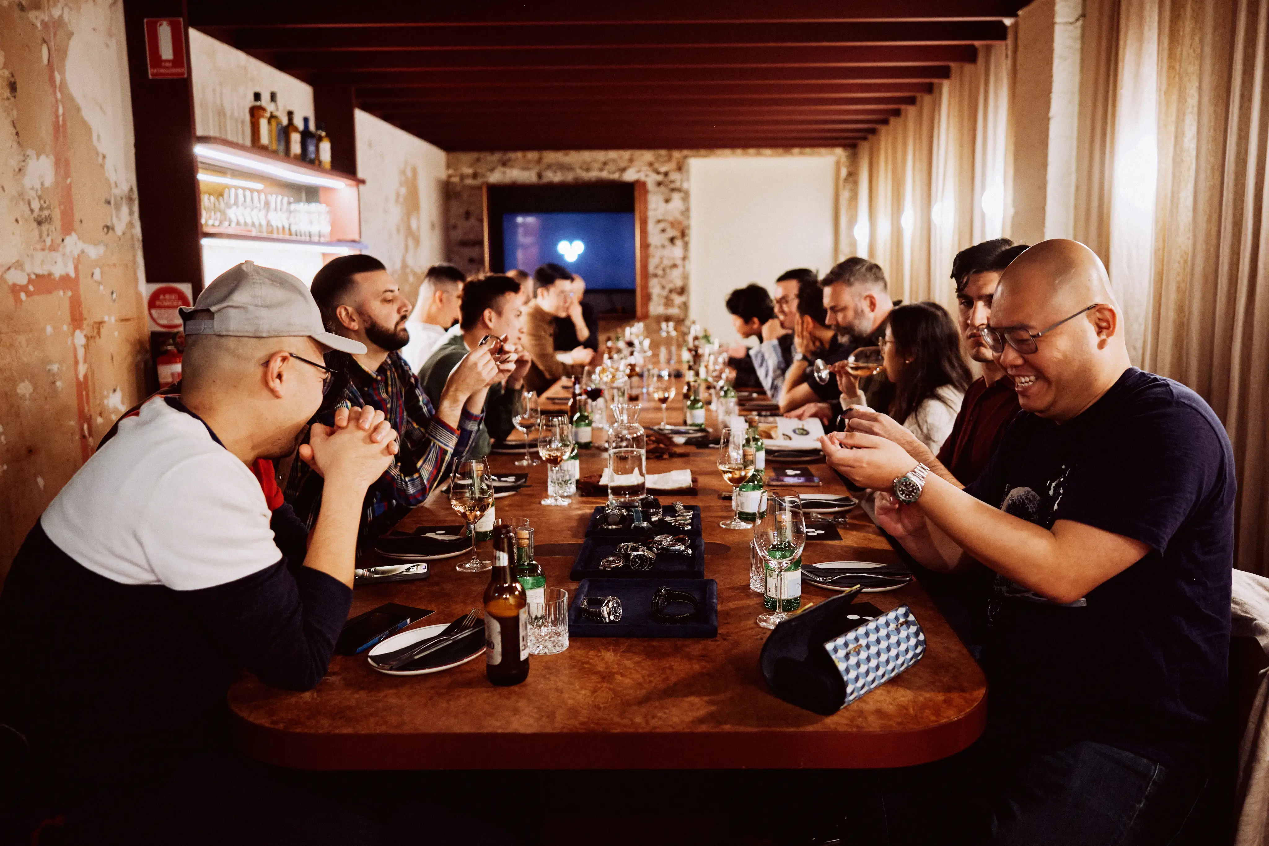 Group of people seated at a long table enjoying drinks and examining wristwatches displayed at the center.