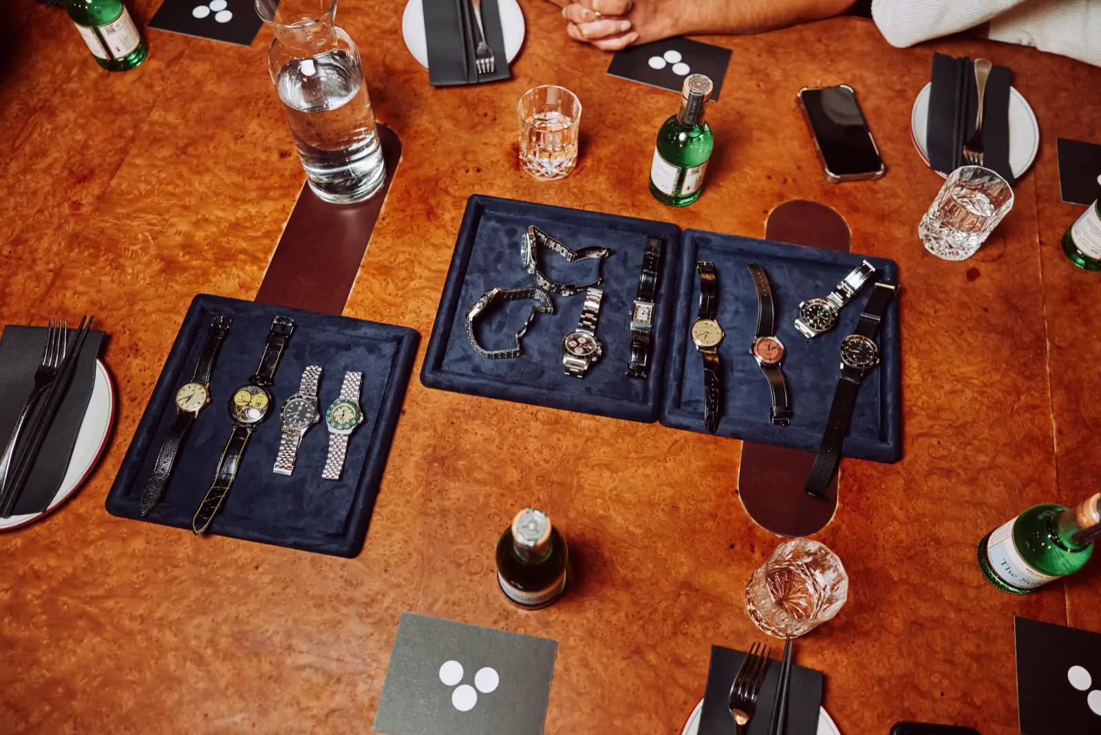 Wooden table with three navy blue trays displaying multiple wristwatches, surrounded by glassware, green bottles, plates, and silverware at a social gathering.