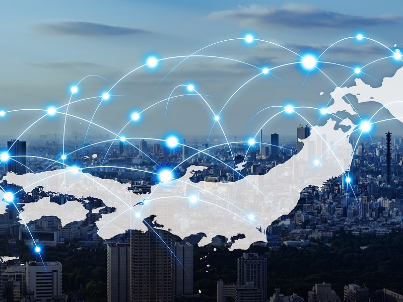 都市の背景に日本地図の輪郭と青い光の接続点が重なり合い、通信ネットワークを表現している。