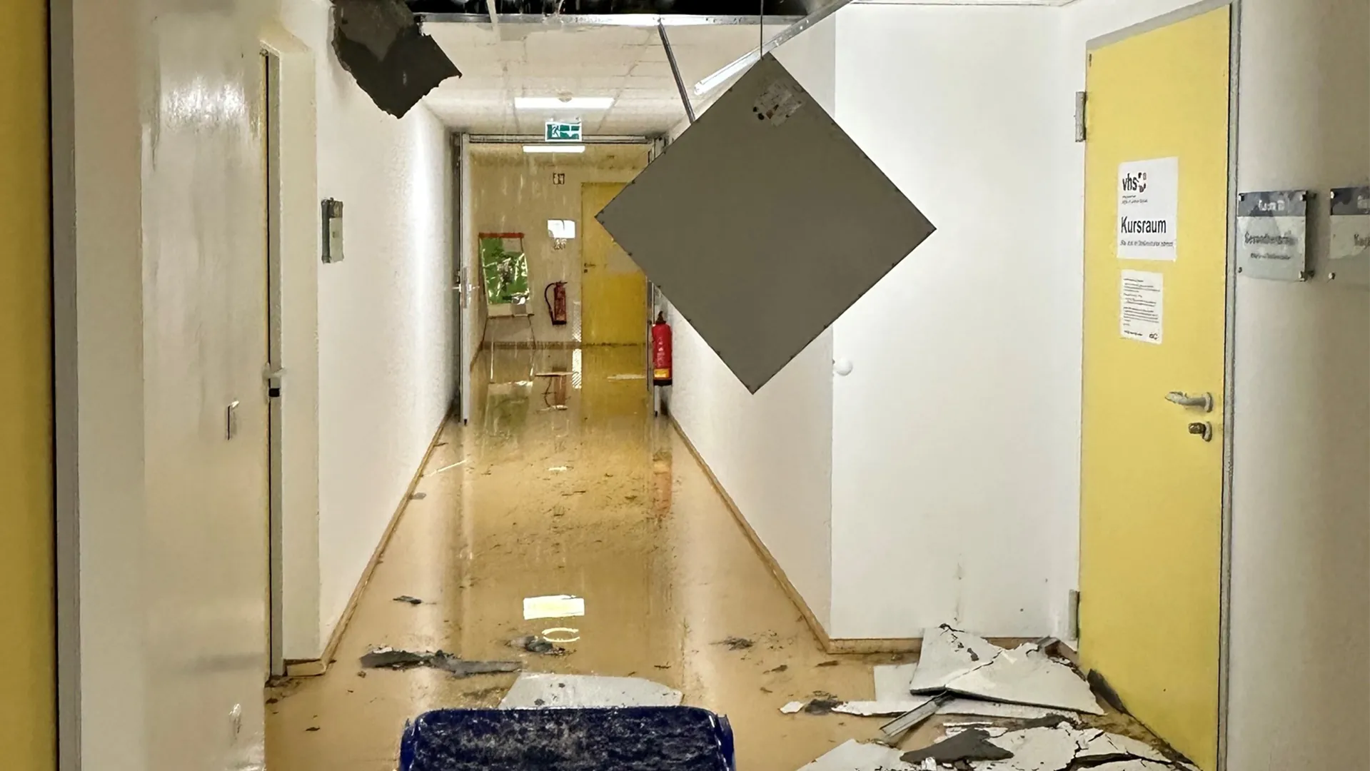 Flur nach Elementarschaden mit beschädigter Decke, herabhängender Deckenplatte und Schutt auf dem Boden, gelbe Tür mit Schild 'Kursraum'