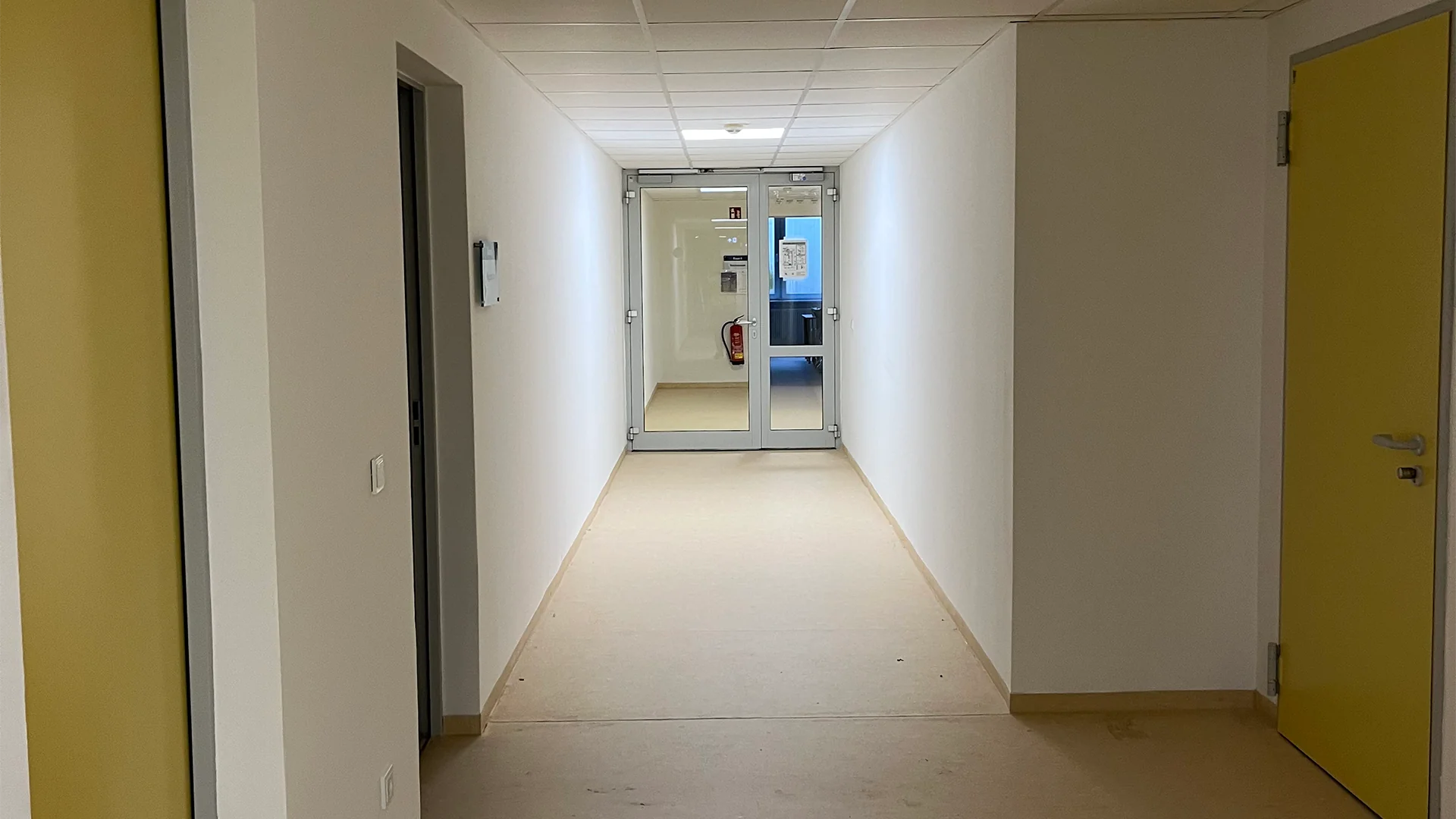 Langer Flur nach der Sanierung mit weißen Wänden, grauem Bodenbelag und mehreren Türen, am Ende eine Glastür mit Blick nach draußen.