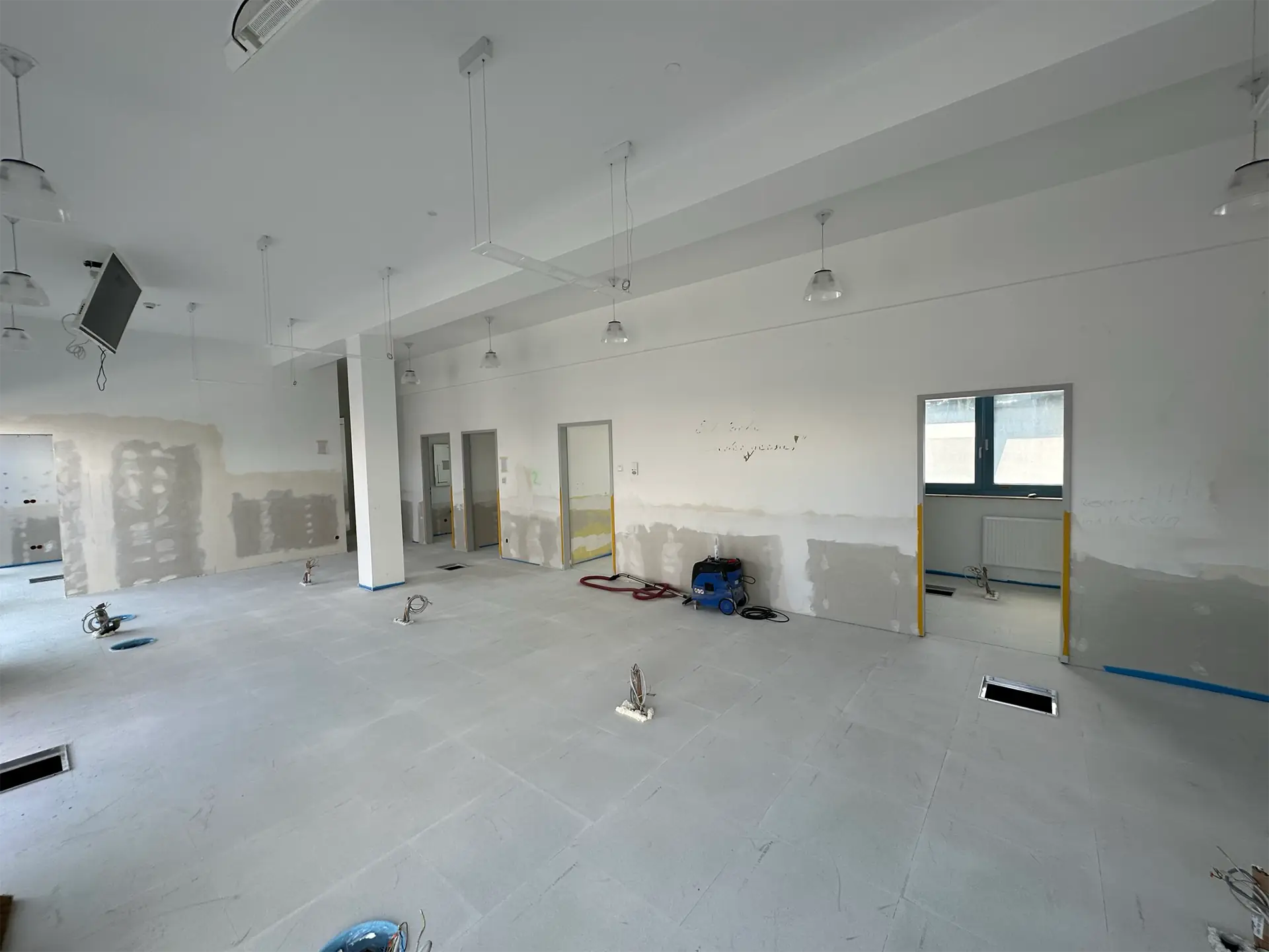 Blick in einen großen, unvollendeten Raum mit grauem Boden. An den Wänden sind unvollständige Putzarbeiten sichtbar. Zwei Türen führen in angrenzende Räume. An der Decke hängen mehrere Lampen. Der Raum befindet sich noch in Sanierung.