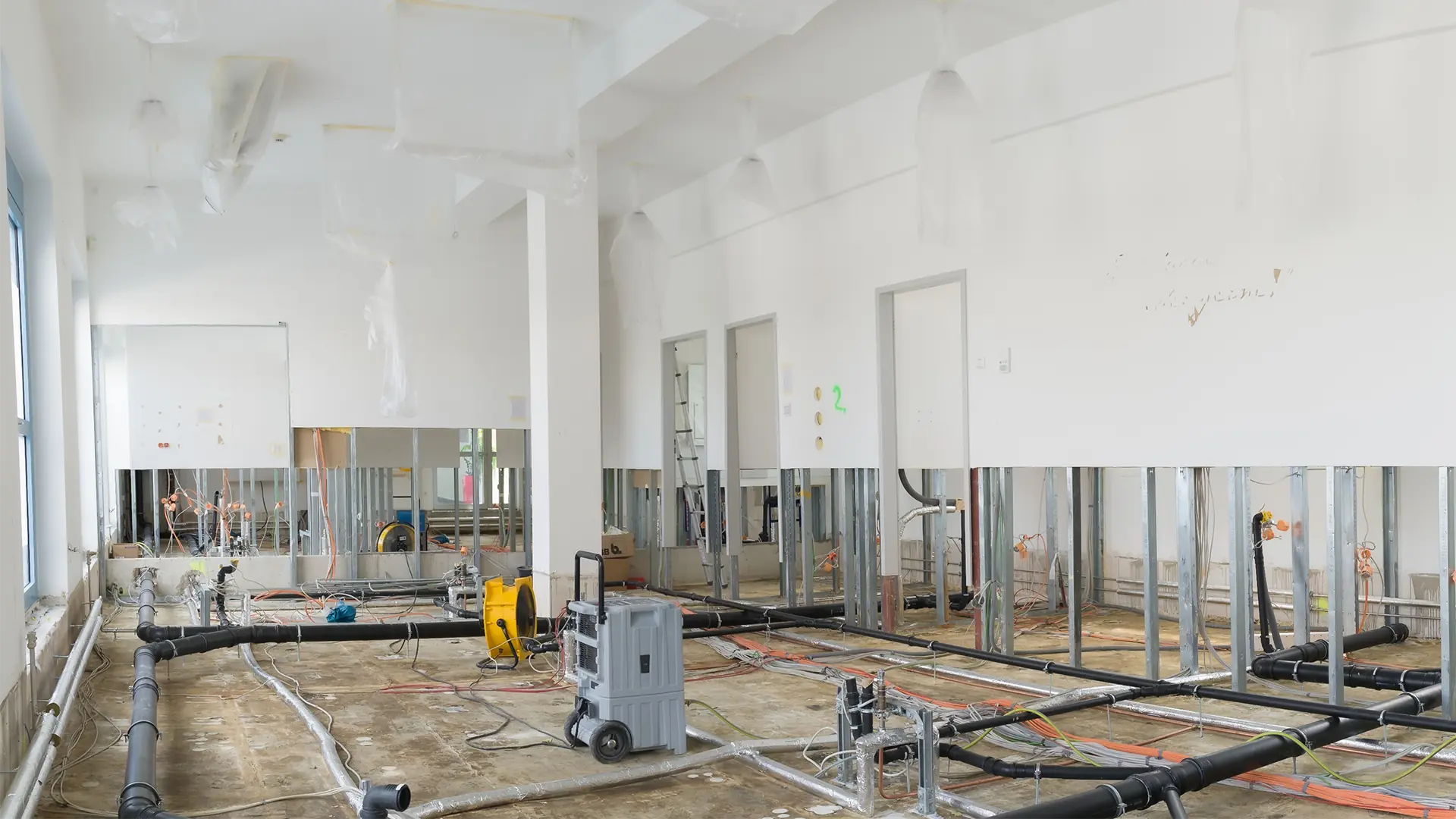 Blick auf einen unfertigen Raum mit freiliegenden Rohren und Kabeln auf dem Boden. An den Wänden sind Metallrahmen für Trockenbau sichtbar. Einige Bereiche sind mit Plastikfolie abgedeckt.