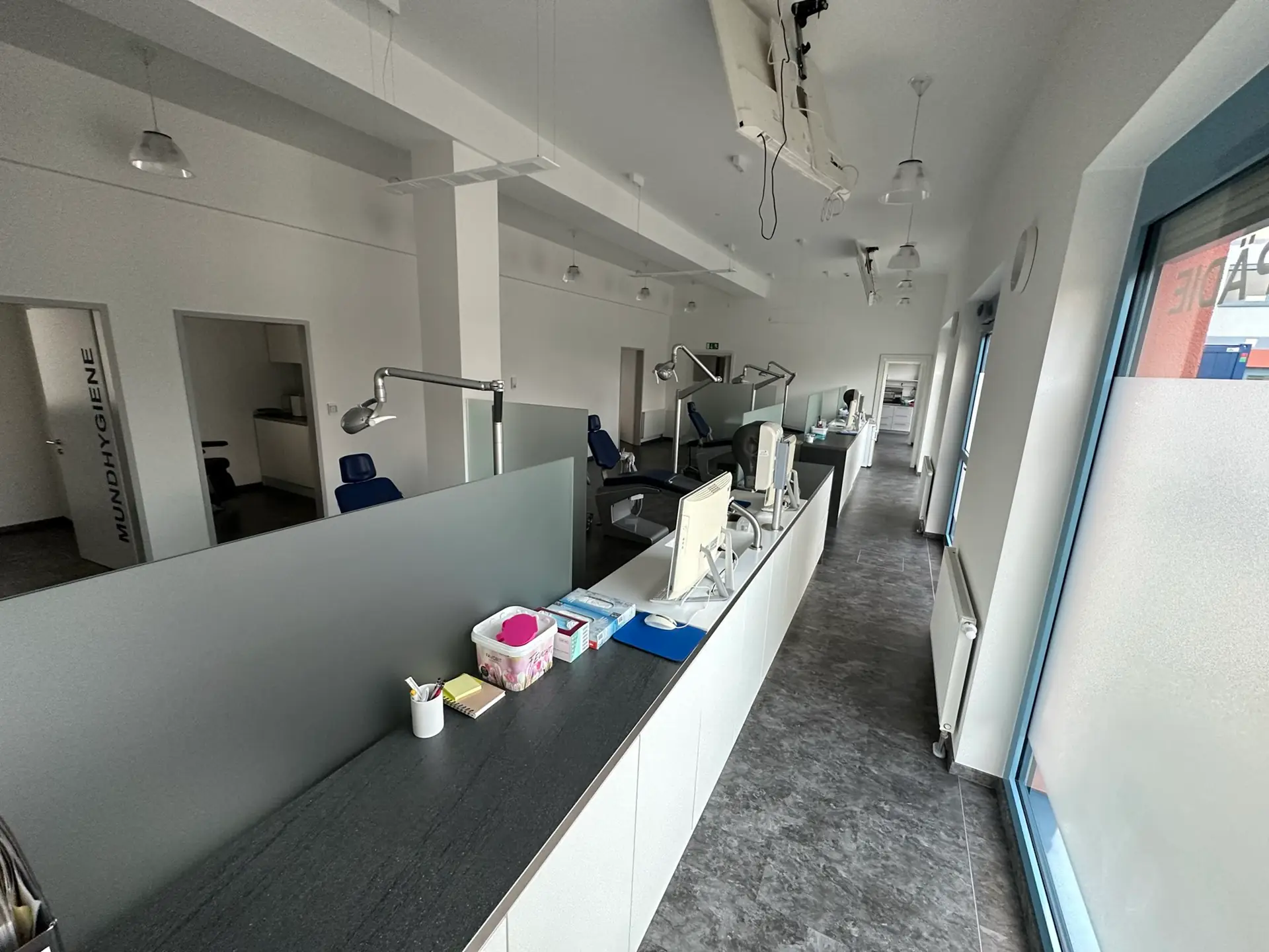 Ein langer Praxisraum mit mehreren Arbeitsstationen nach Sanierungsarbeiten. Im Vordergrund sind Utensilien und Monitore sichtbar. Die Arbeitsstationen sind durch Milchglaswände getrennt.