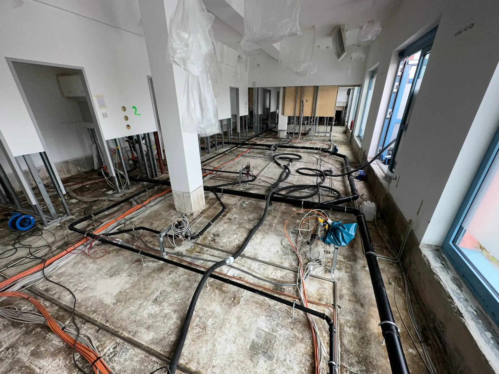 Blick auf einen unfertigen Raum mit freiliegenden Rohren und Kabeln auf dem Boden. An den Wänden sind Metallrahmen für Trockenbau sichtbar. Einige Bereiche sind mit Plastikfolie abgedeckt.