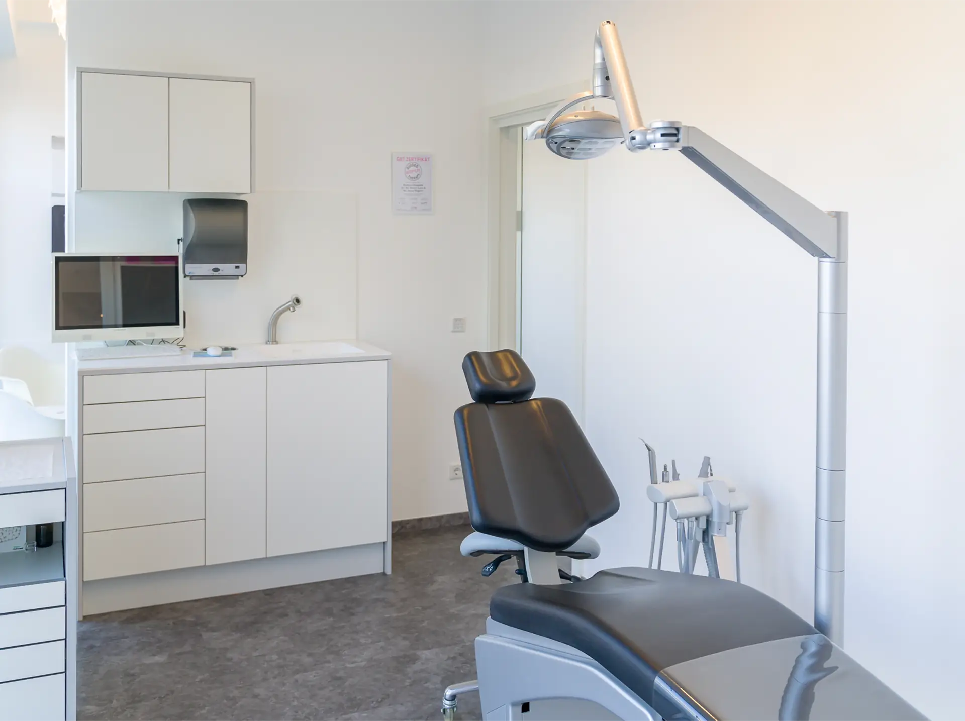 Behandlungszimmer nach den abgeschlossenen Sanierungsarbeiten. Im Vordergrund ist ein schwarzer Behandlungsstuhl, daneben eine einstellbare Leuchte. Im Hintergrund sind weiße Schränke, ein Computer und Sanitäranlagen zu sehen.