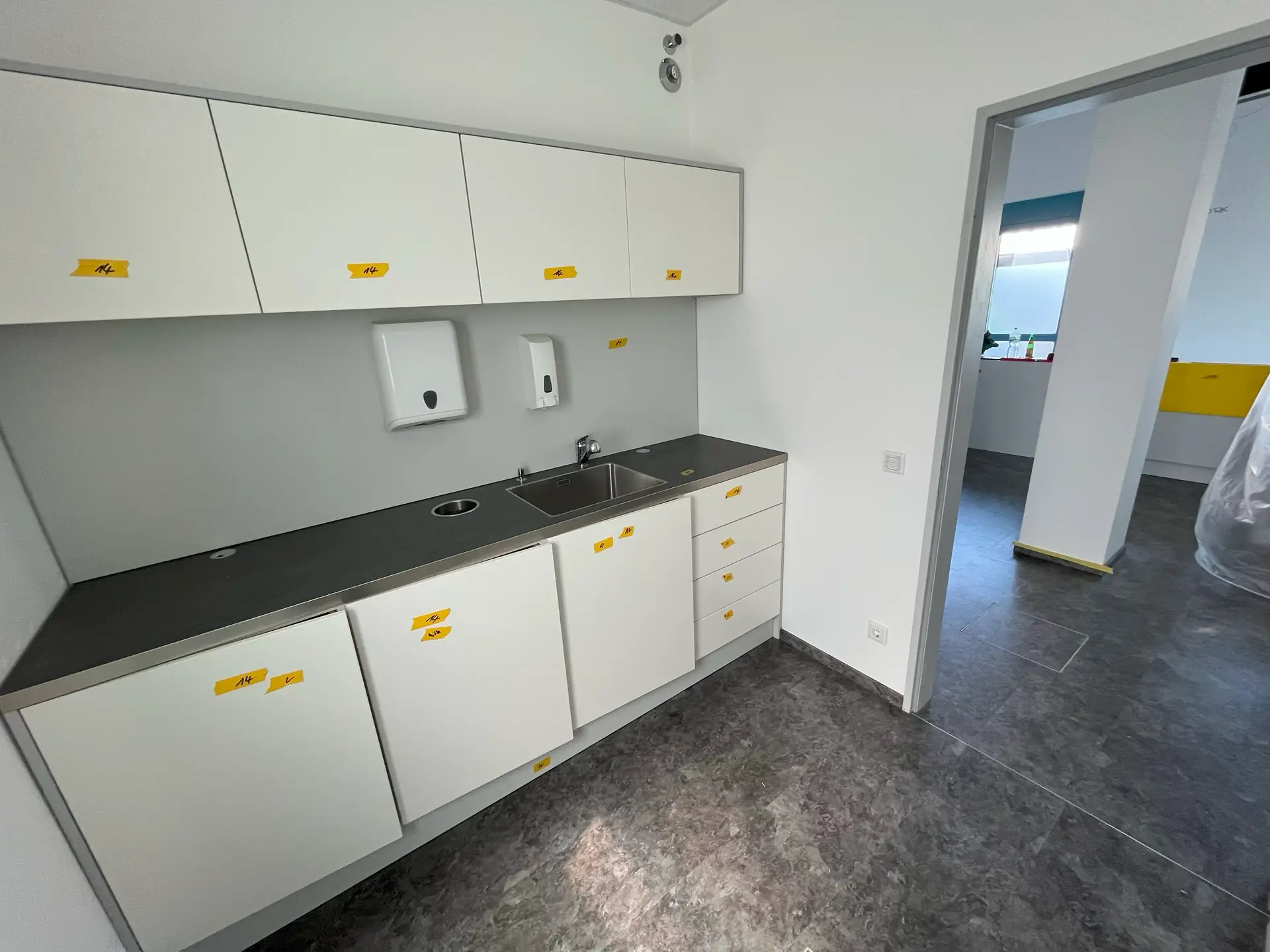 Küchenzeile mit weißen Schränken, grauer Arbeitsplatte und Spüle nach Sanierungs- und Renovierungsarbeiten. Zwei Seifenspender an der Wand. Blick auf einen angrenzenden Raum.