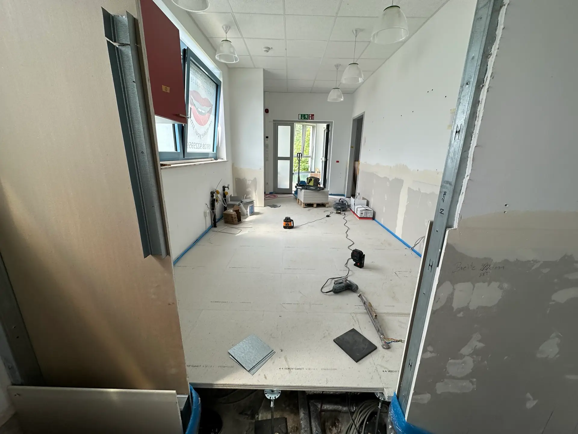 Blick in einen unfertigen Raum mit einem hellen Bodenbelag. Auf dem Boden liegen verschiedene Werkzeuge, darunter ein Staubsauger und Platten. An der Wand sind unvollständige Verkleidungen zu sehen. Der Raum befindet sich gerade in Sanierung.
