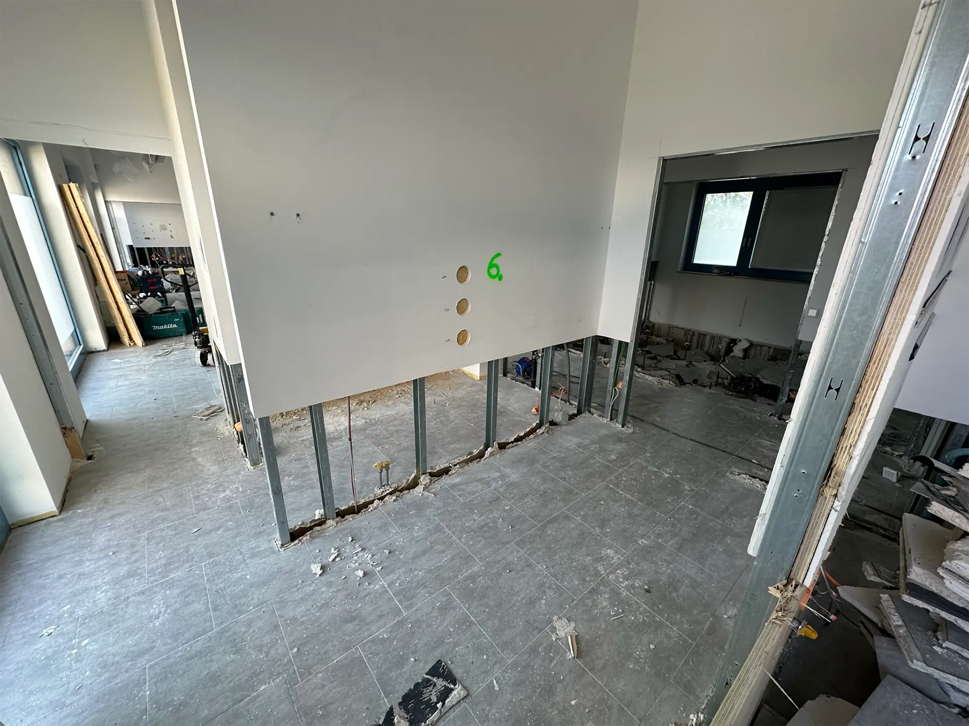Blick auf einen Raum mit noch nicht beendeten Bauausführung. An einer Wand sind sechs runde Löcher sichtbar, der Boden ist teilweise abgedeckt und es liegen Bauabfälle herum.