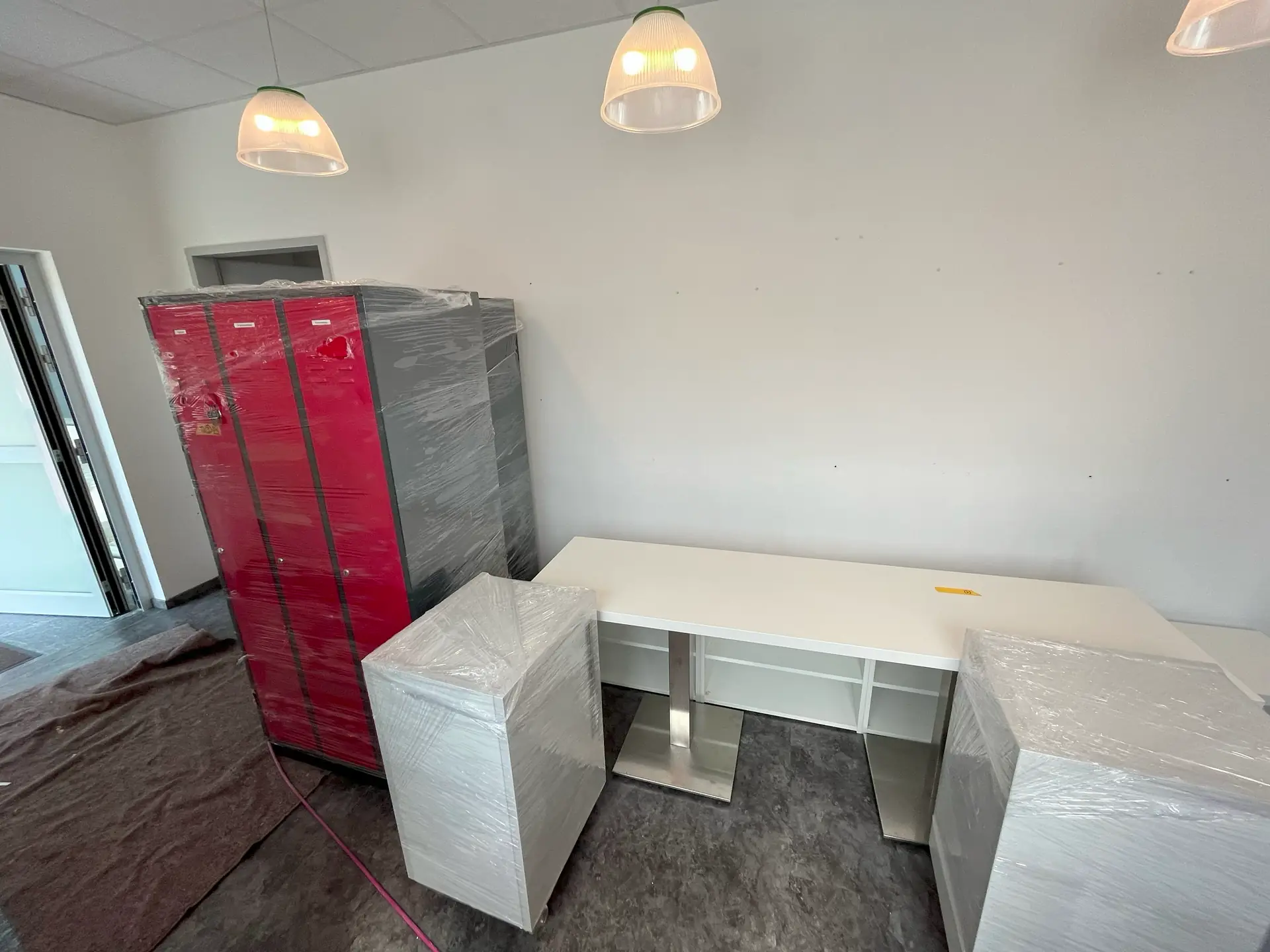 Ein Raum mit drei roten Schränken, die in Folie eingewickelt sind, und einem weißen Tisch mit zwei weißen Stühlen, ebenfalls eingewickelt. Deckenleuchten sind sichtbar.