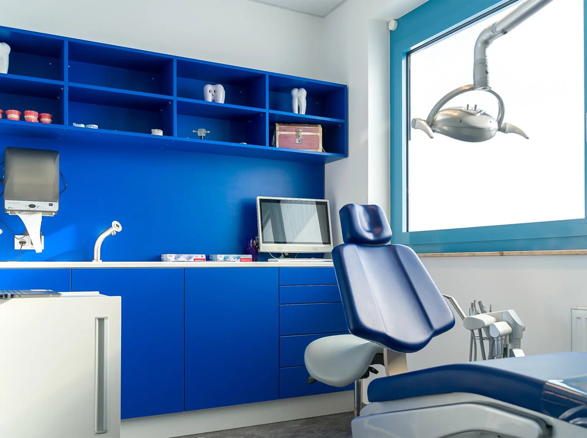 Behandlungszimmer nach den abgeschlossenen Sanierungsarbeiten. Im Vordergrund ist ein blauer Behandlungsstuhl, daneben eine einstellbare Leuchte. Im Hintergrund sind leuchtend blaue Schränke, ein Computer und Sanitäranlagen zu sehen. Ein großes Fenster füllt den Raum mit Licht.