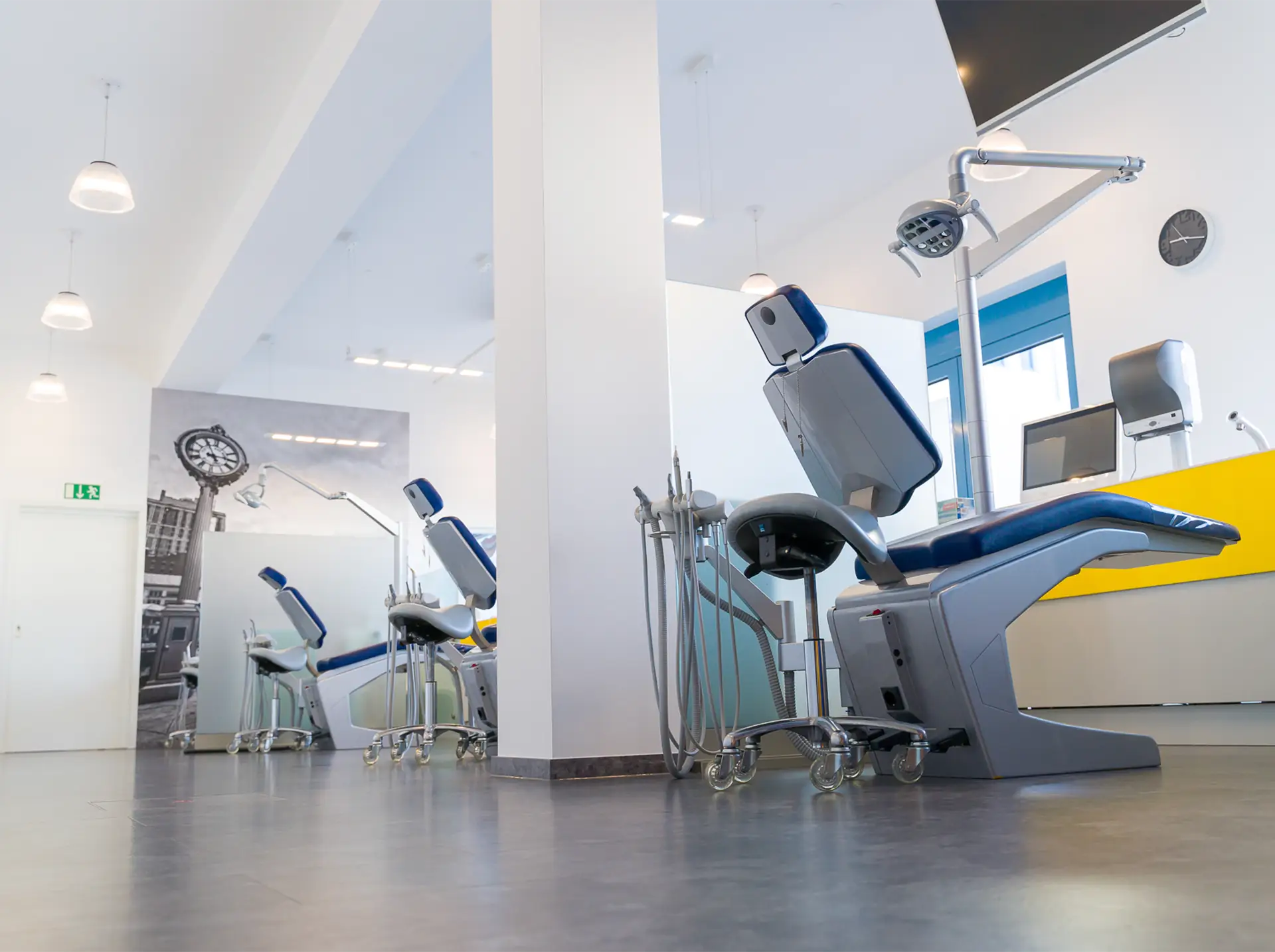 Fertig sanierte Praxisräume mit mehreren Behandlungssektionen, die durch undurchsichtige Glaswände getrennt sind. Der Raum ist durch Deckenleuchten und große Fenster hell erleucht. Im Vordergrund sind die Behandlungsstühle und -lichter zu sehen.