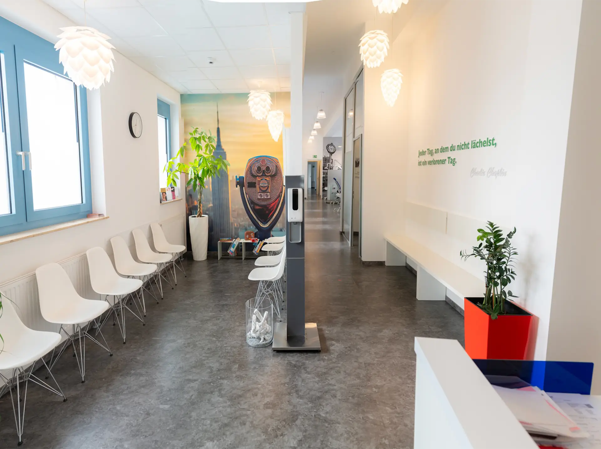 Fertiger Eingangsbereich einer Zahnarztpraxis nach Sanierungsarbeiten. Auf der linken Seite ist der Wartebereich mit zehn weißen Stühlen und einer grünen Pflanze zu sehen, auf der rechten Seite ist ein Flur mit einer weißen Bank und einem Schriftzug an der rechten Wand zu sehen. Beide Bereiche werden durch eine dünne Mittelwand getrennt, an der ein Desinfektionsmittelspender steht.