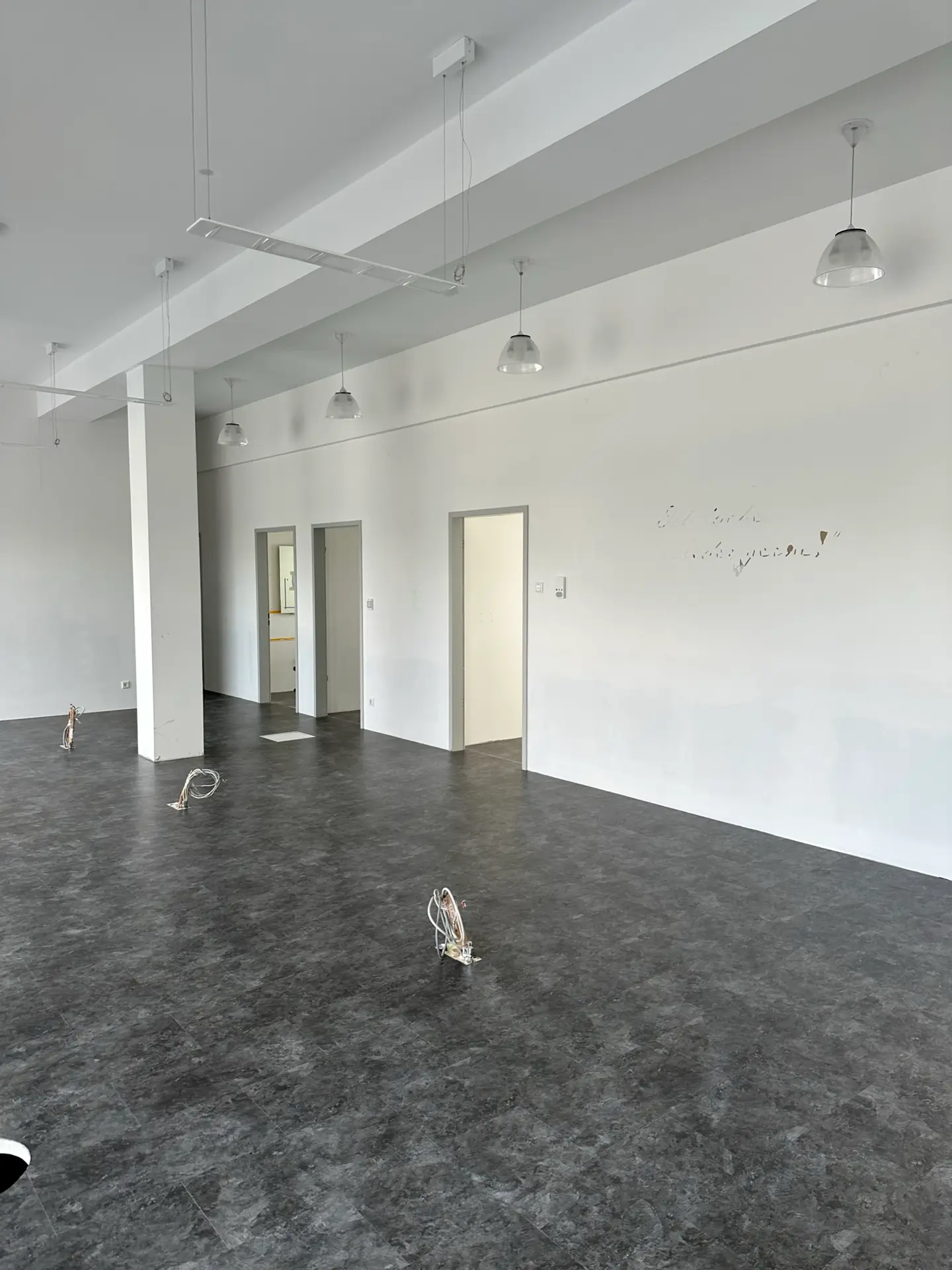 Leerer Raum mit grauem Boden und weißen Wänden. An der Decke hängen mehrere Lampen. Zwei Türen sind sichtbar, eine Wand hat handschriftlichen Text. Die Sanierungs- und Renovierungsarbeiten sind fast abgeschlossen.