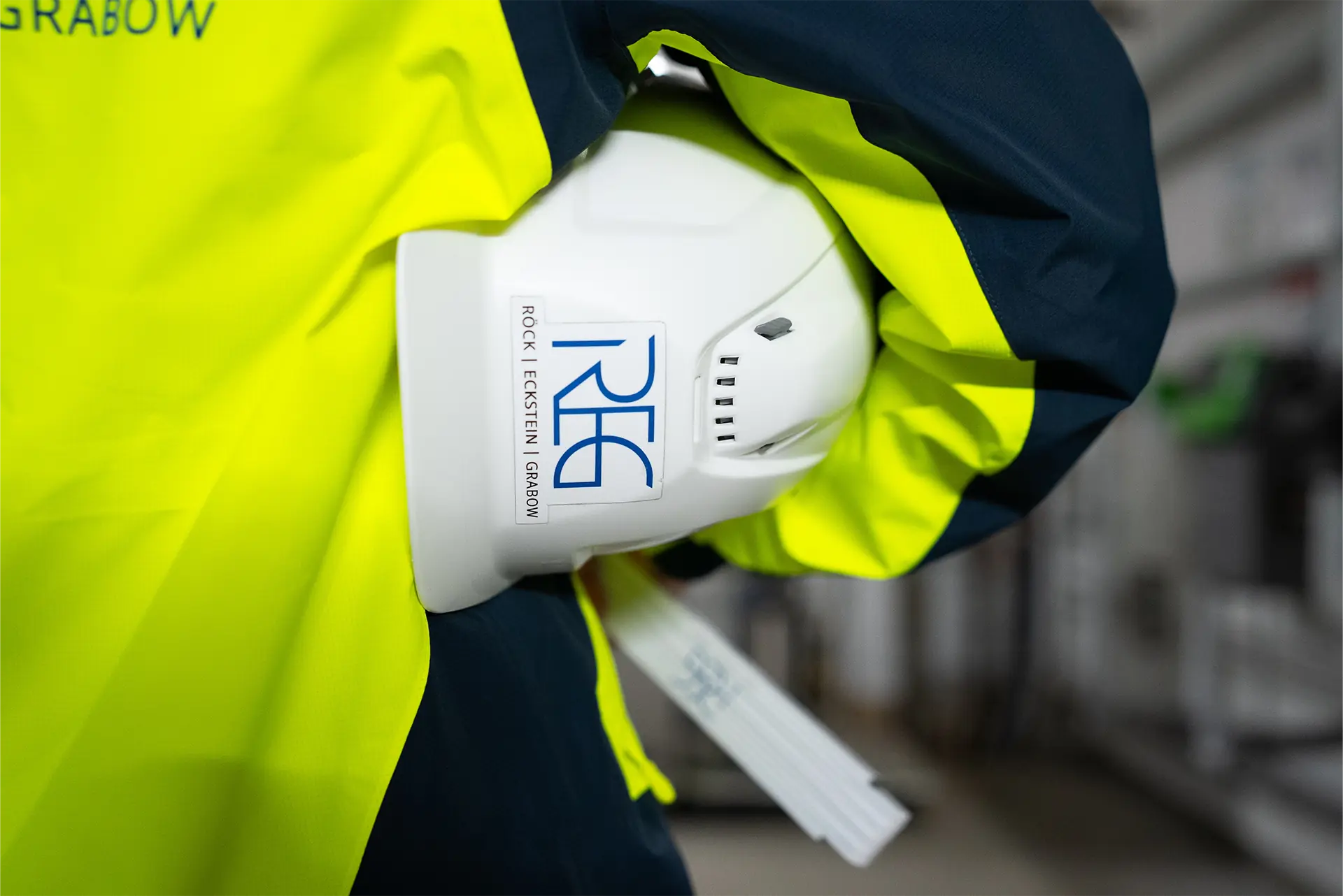 Eine Person trägt eine gelbe Warnjacke und hält einen weißen Helm unter dem rechten Arm geklemmt. In der rechten Hand hält sie einen Meterstab.