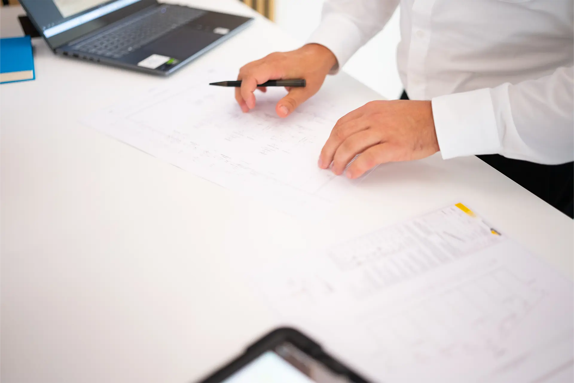 Ein Mann mit einem weißen Hemd hält einen Stift über Bauplänen auf einem Tisch. Im Hintergrund sind ein Laptop und ein Notizbuch sichtbar.