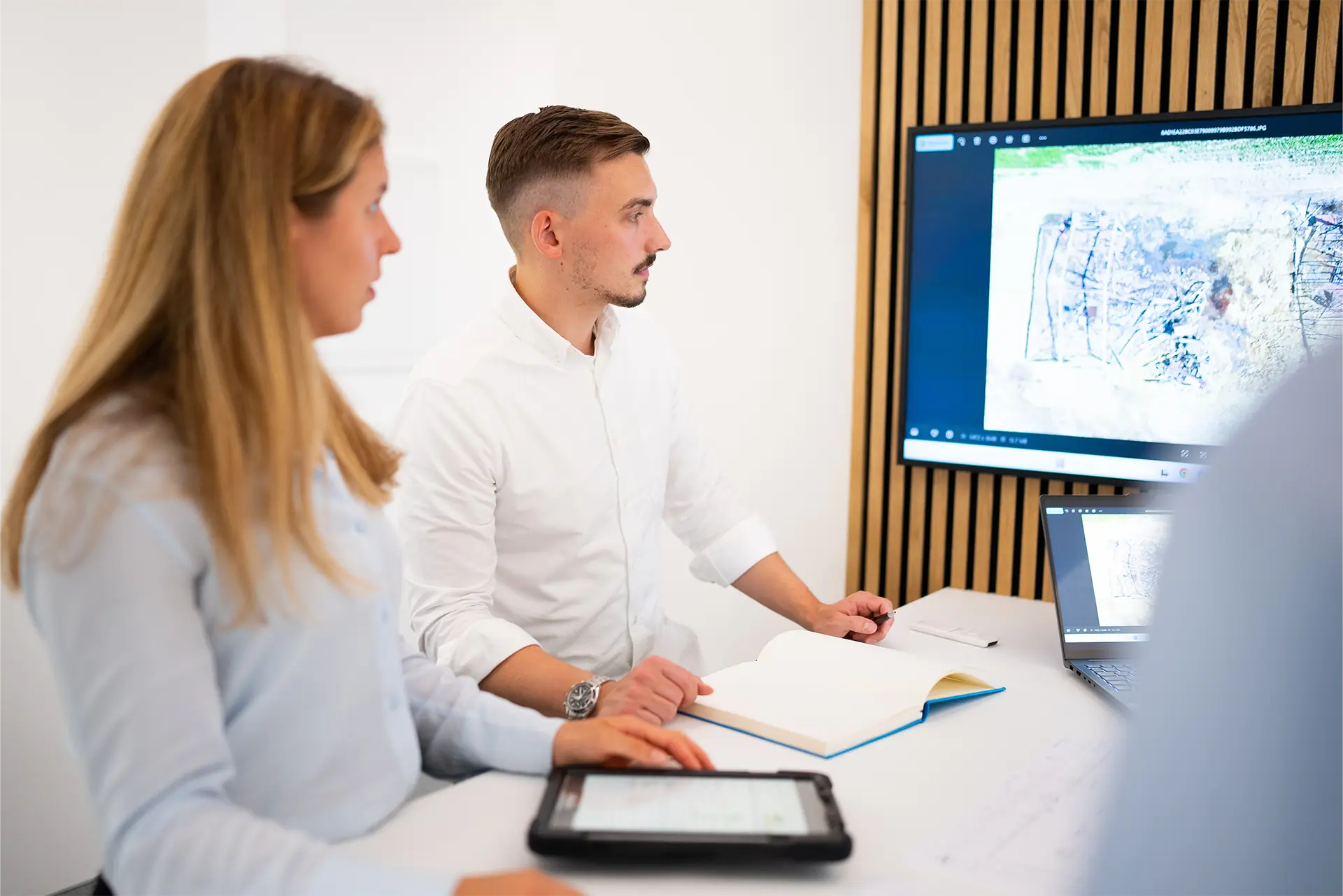 Drei Personen in einem Besprechungsraum, eine Person steht am Tisch mit einem geöffneten Notizbuch. Auf dem Tisch liegt ein Tablet und ein Monitor zeigt eine Karte.