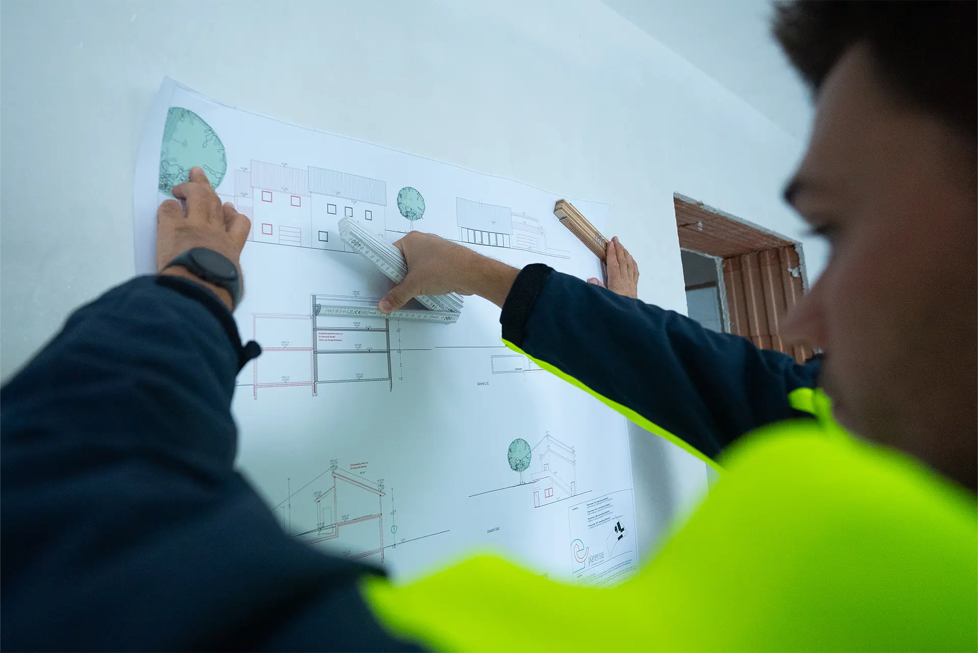 Eine Person in einer gelben Sicherheitsweste hält einen Bauplan an einer Wand. Die Person benutzt ein Lineal, um Maße auf dem Plan zu überprüfen.