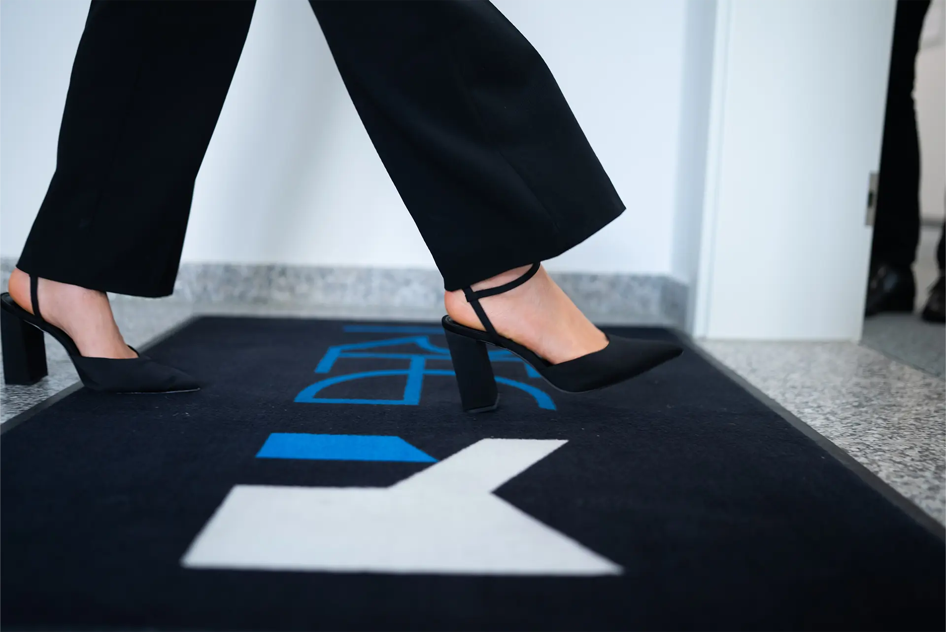 Eine Person mit schwarzen High Heels und schwarzen Hosen geht über einen Teppich mit einem blauen Logo von REG und einem weiß-blauen Logo eines "M".
