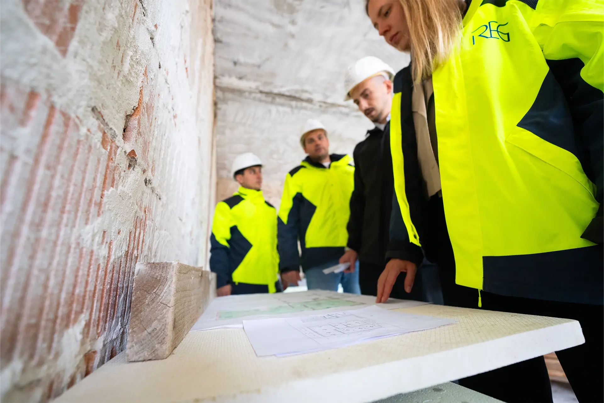 Gruppe von Personen in leuchtend gelben Sicherheitsjacken mit weißen Helmen, die über einen Tisch mit Plänen und Materialien in einem Raum mit Ziegelwänden beugen.
