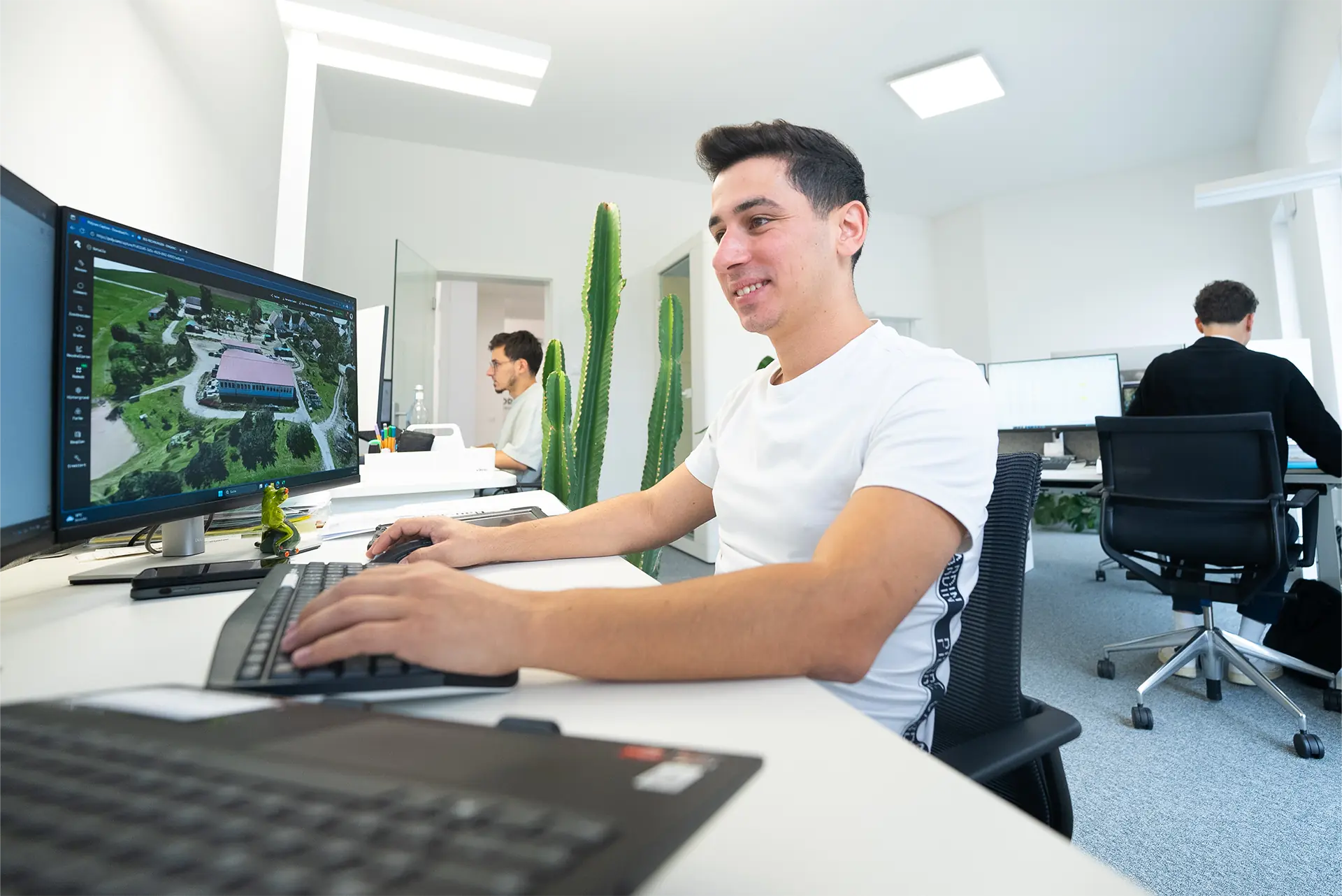 Mann sitzt an einem Schreibtisch mit Computer, arbeitet mit einer Software. Im Hintergrund sind zwei weitere Personen und Pflanzen sichtbar. Der Mann trägt ein weißes T-Shirt.