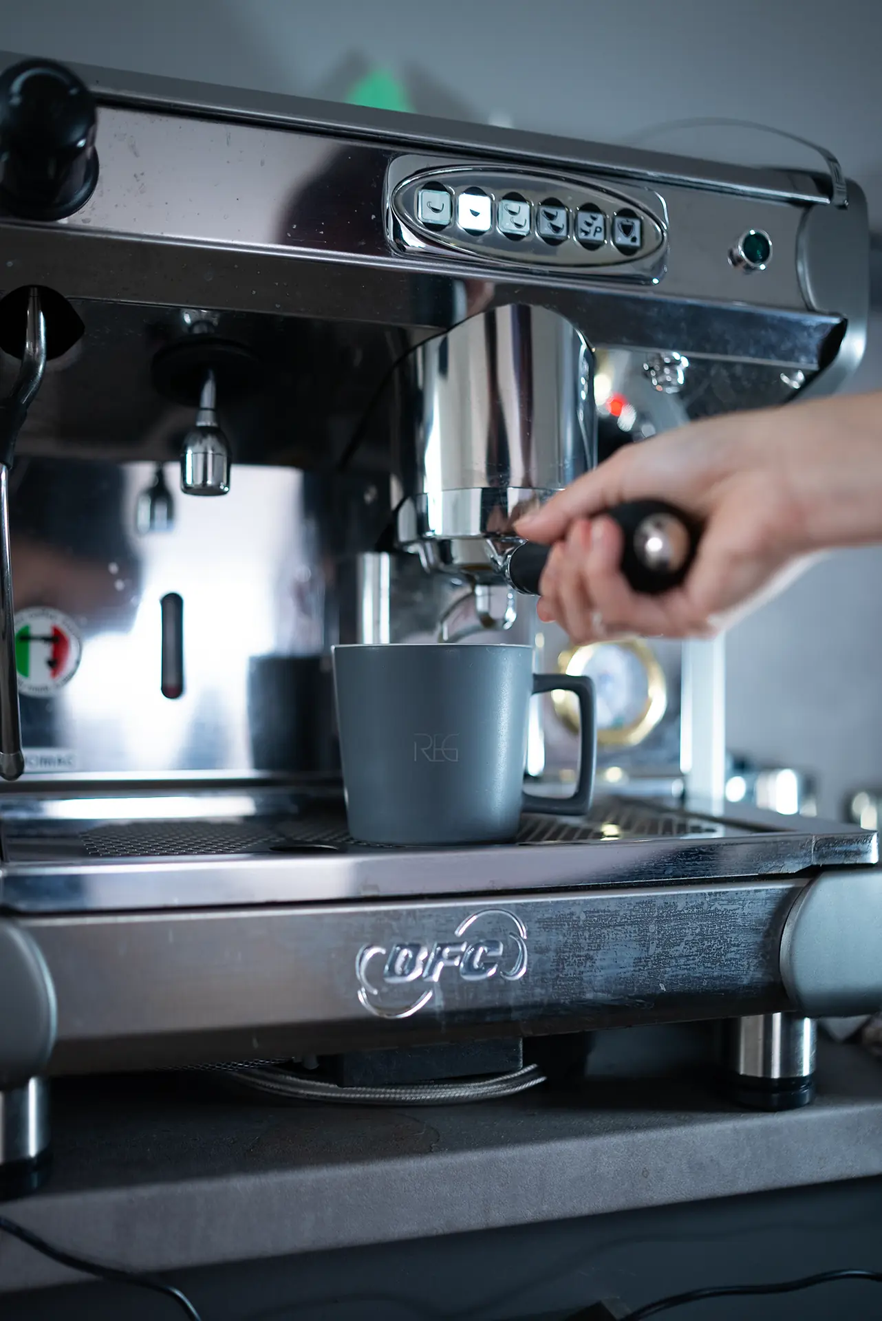 Eine Hand hält einen schwarzen Griff eines Siebträgers einer Kaffeemaschine, während ein Espresso in eine graue Tasse fließt. Die Maschine hat eine glänzende Metalloberfläche.