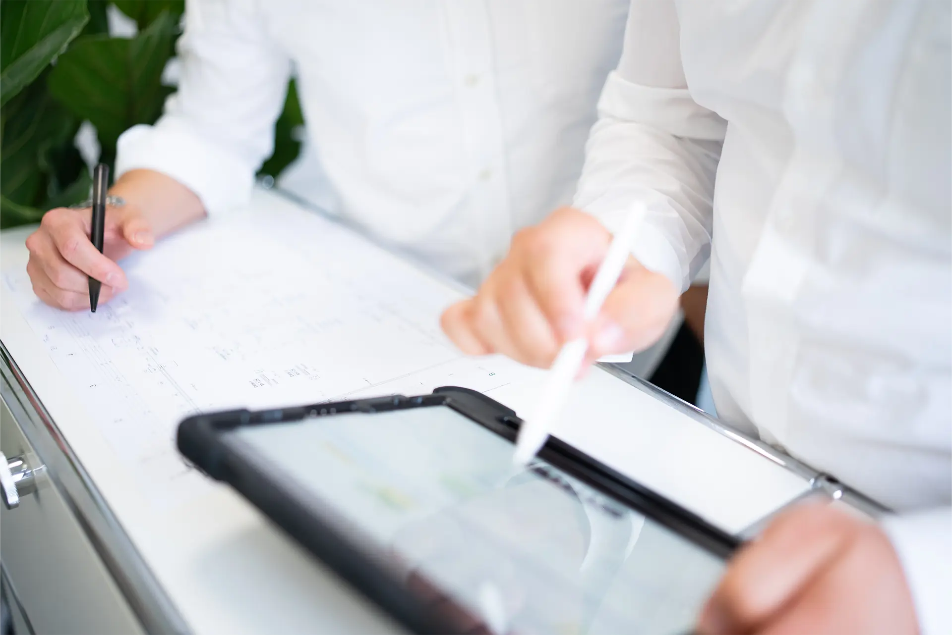 Zwei Personen in weißen Hemden arbeiten an einem Tisch. Eine Person hält einen Stift und macht Notizen auf einem Blatt Papier, während die andere Person einem Tablet mit einem Stylus bedient.