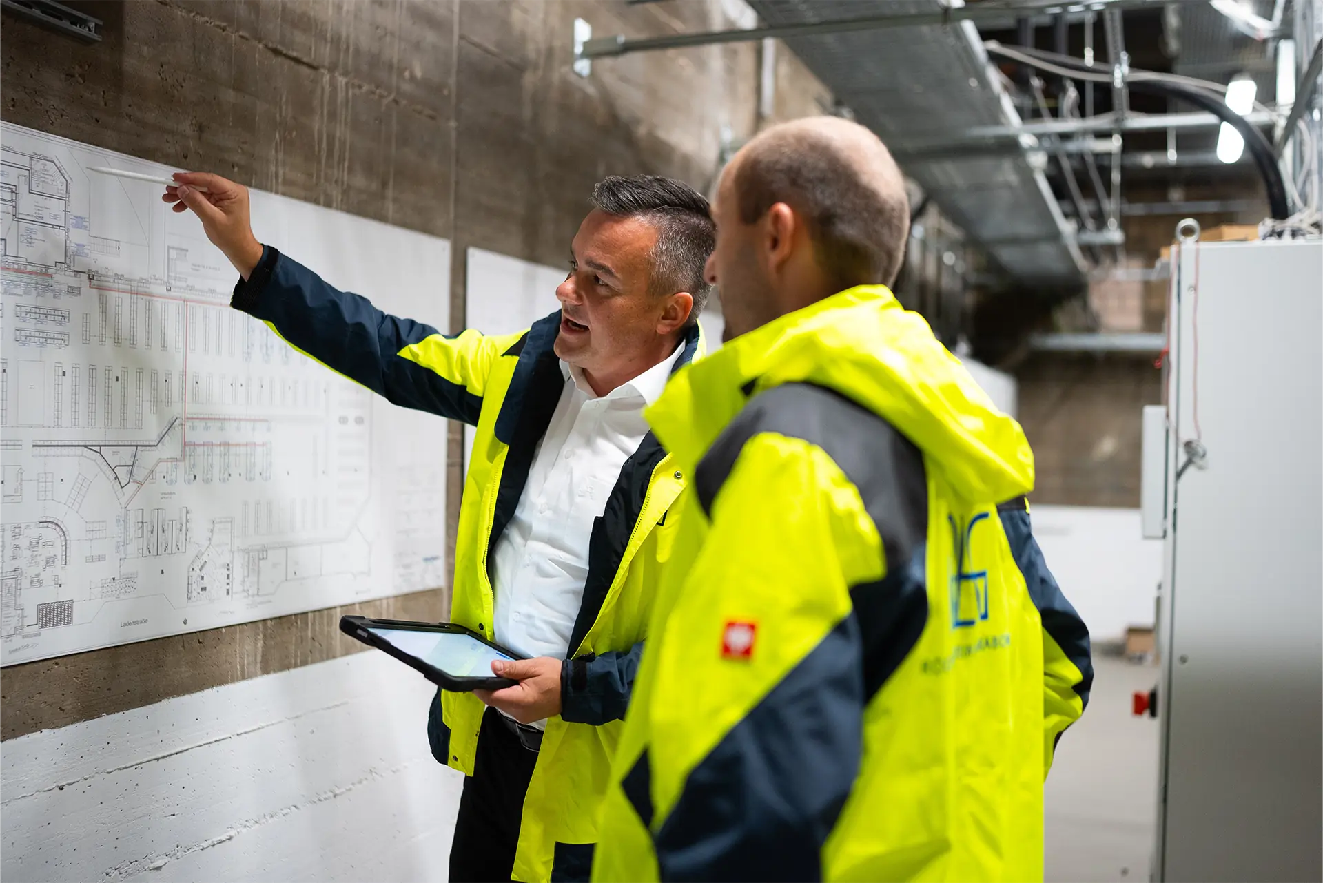Zwei Personen in gelben Sicherheitsjacken stehen vor einer Wand mit einem Bauplan. Eine Person zeigt mit dem Finger auf den Plan und hält dabei ein Tablet in der anderen Hand.