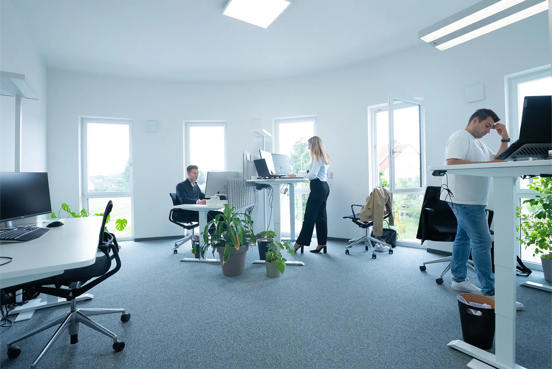 Büro mit drei Personen: eine Frau steht am Schreibtisch, ein Mann sitzt an einem Computer, ein weiterer Mann steht nachdenklich an seinem Arbeitsplatz. Pflanzen und moderne Möbel sind sichtbar.