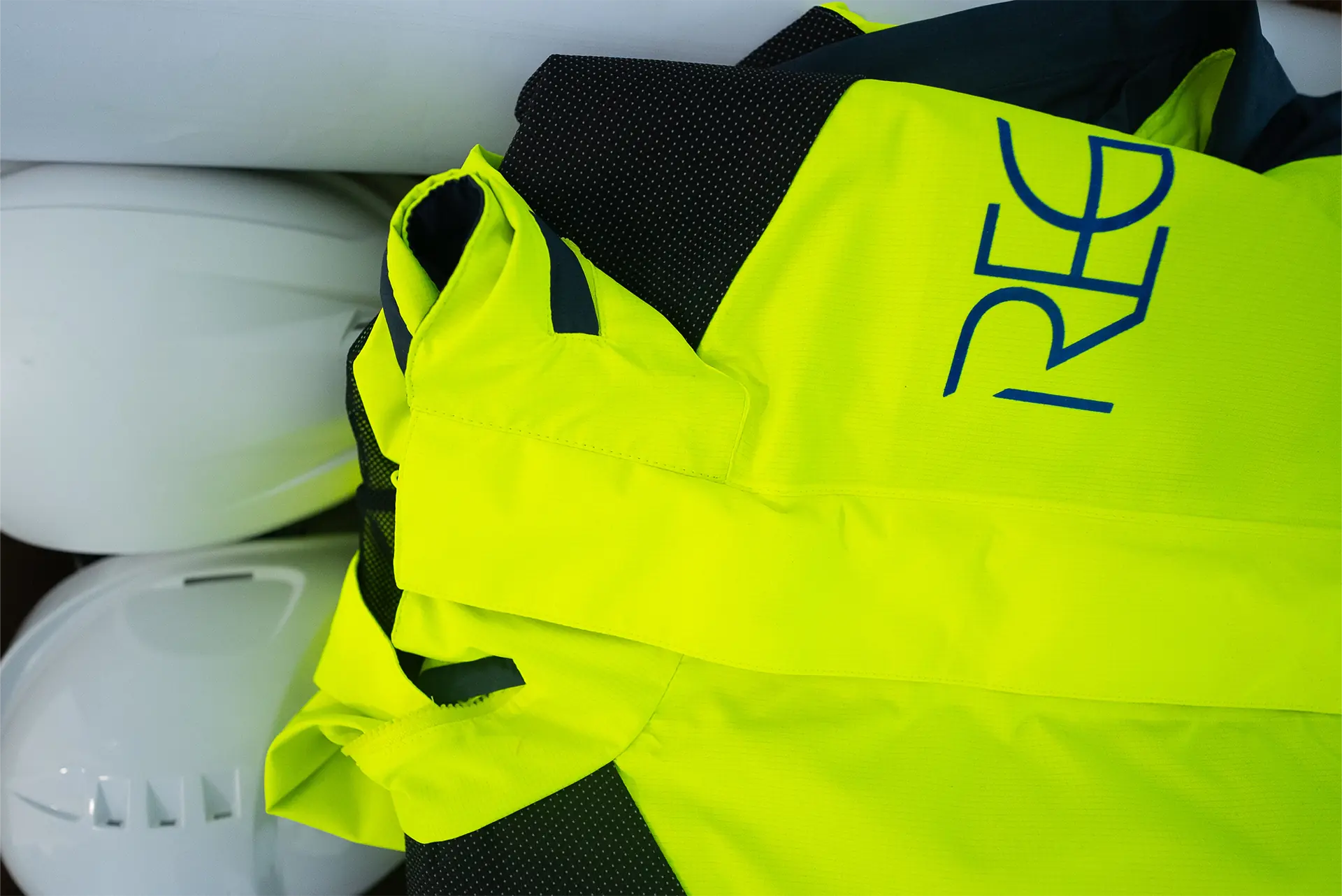Zusammengelegte, gelbe Sicherheitsjacke mit schwarzem Netzstoff und blauer Schrift 'REG', liegt neben zwei weißen Bauhelmen.