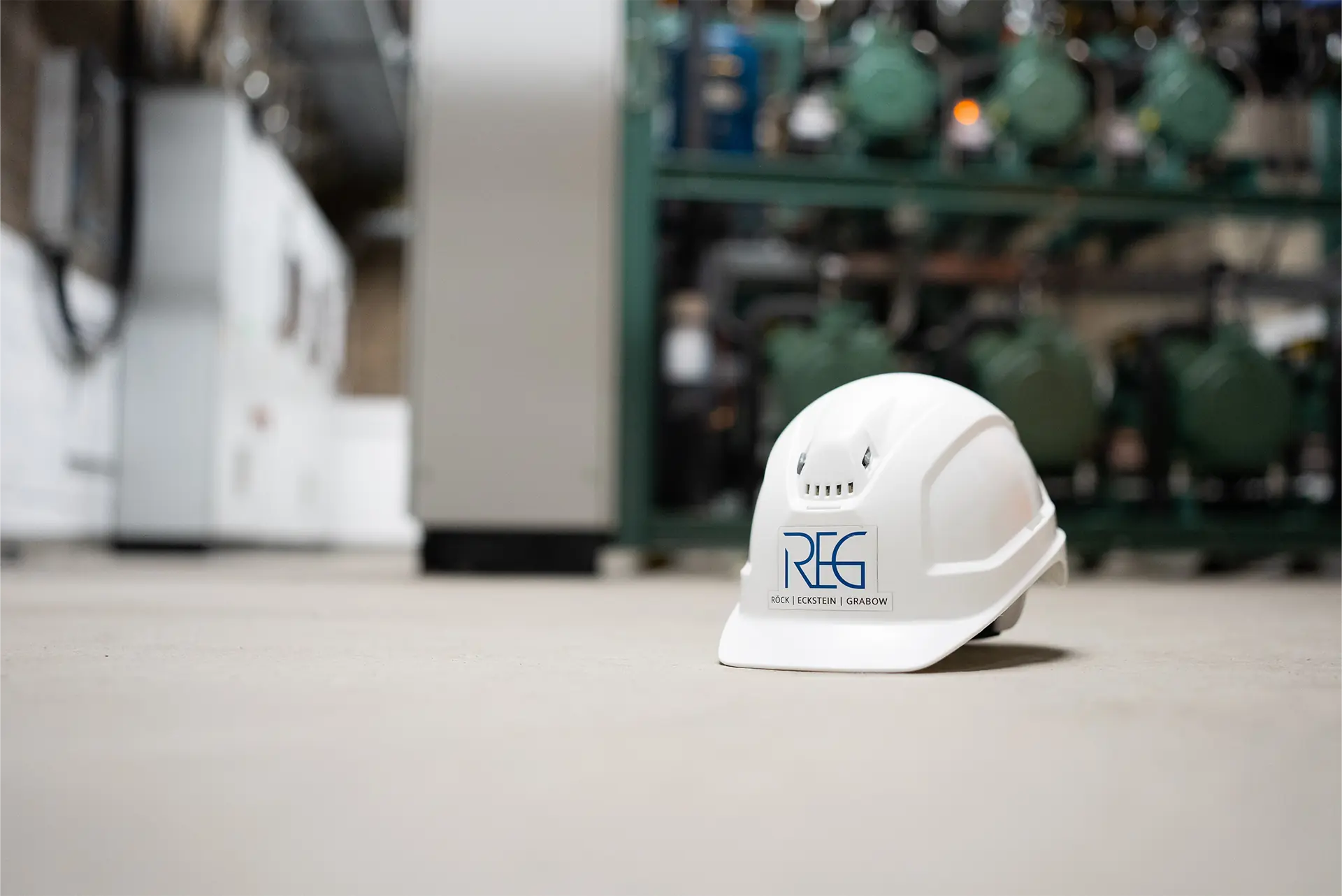 Weißer Sicherheitshelm mit blauer Schrift 'REG' im Vordergrund, im Hintergrund Maschinen und Ausrüstung.