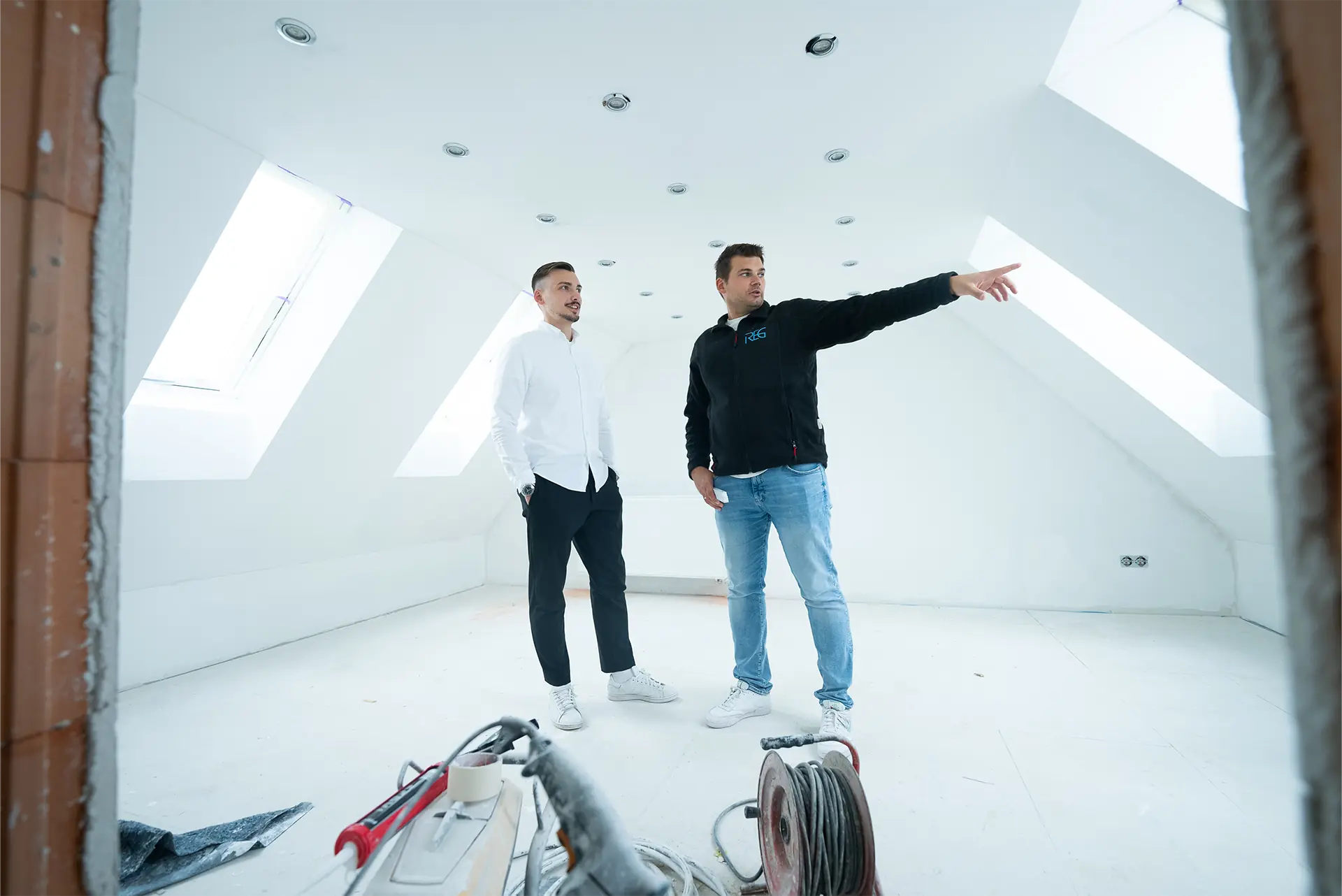 Zwei Personen stehen in einem hellen, leeren, frisch sanierten Raum mit Dachfenstern. Eine Person trägt ein weißes Hemd und die andere ein schwarzes Sweatshirt. Im Vordergrund liegt ein Baugerät.